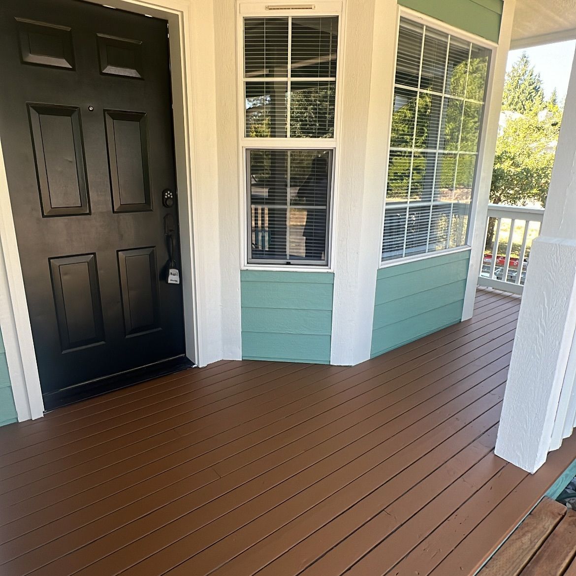 Brown wood porch with a black door, teal siding, and large windows.
