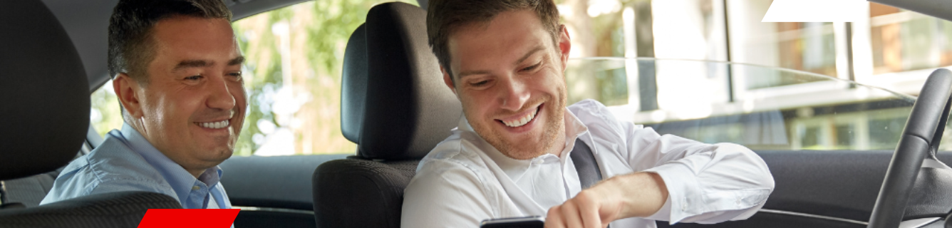 Dois homens sorrindo dentro de um carro, um deles olhando para o celular.