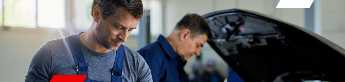 Dois mecânicos de macacão azul examinam o motor de um carro em uma garagem.