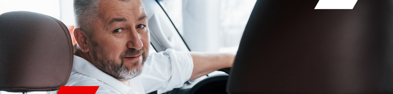 Homem de camisa branca, olhando para a câmera, dentro de um carro, com o banco marrom visível.