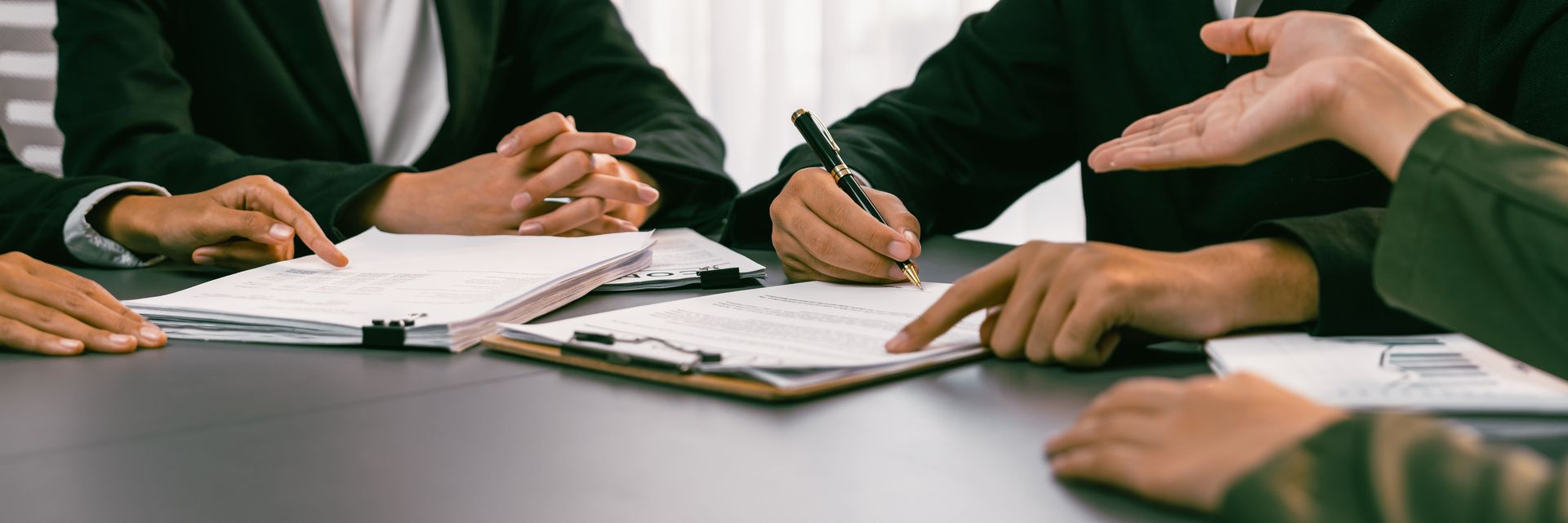 People in suits at a table, some pointing at papers, others writing, in a meeting.