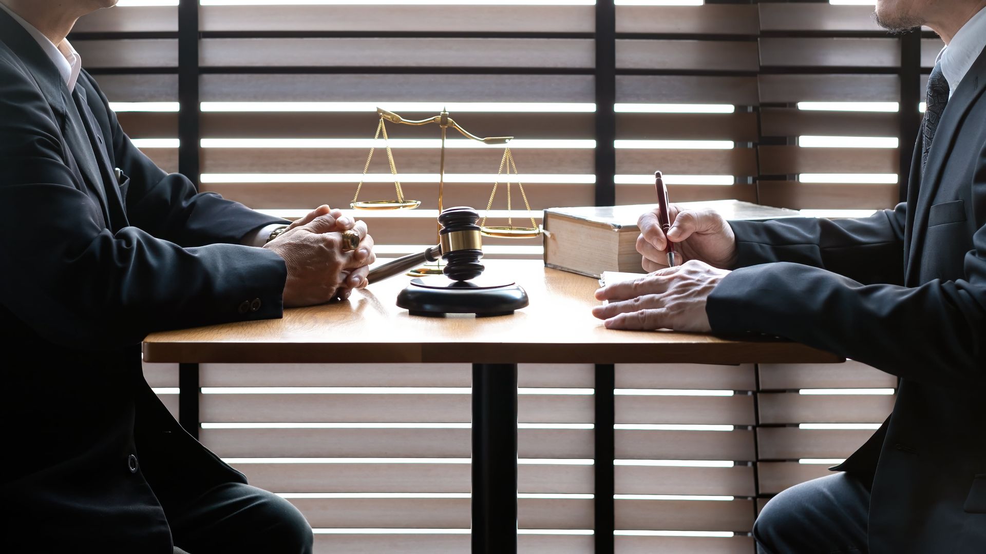 Two people in suits at a table with a gavel, scales, and law books, in front of a window.