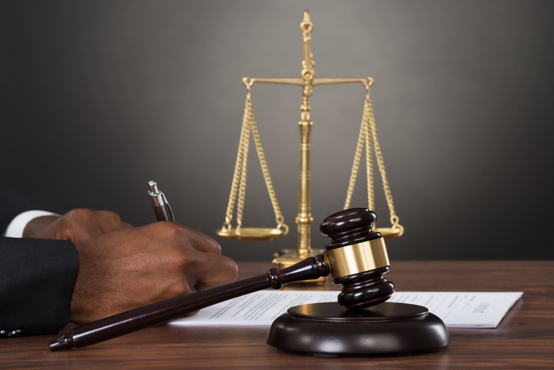 Judge's hand writing on a document with gavel, scales of justice, on a desk.