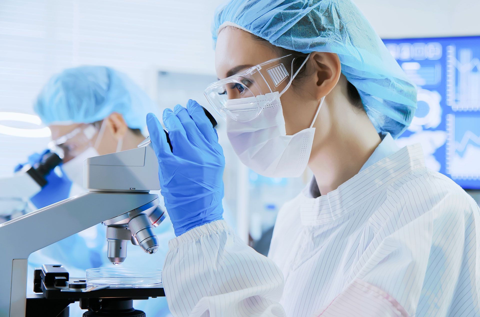 Two lab workers in protective gear examining samples under microscopes in a lab.