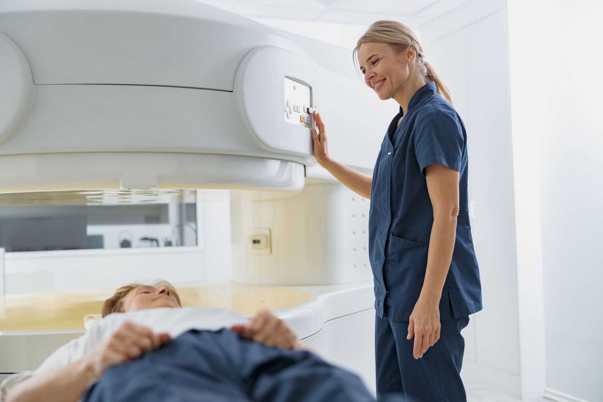 A medical professional smiles while adjusting controls of an MRI machine over a patient.