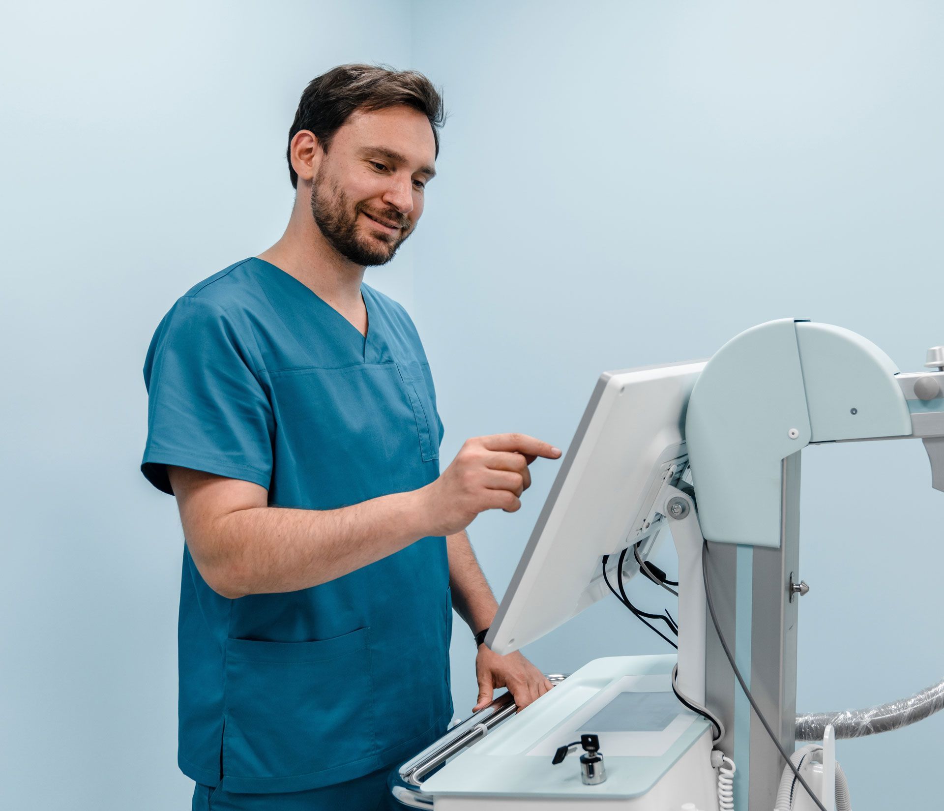 Doctor in blue scrubs smiling, touching a touchscreen display of medical equipment. Light blue background.