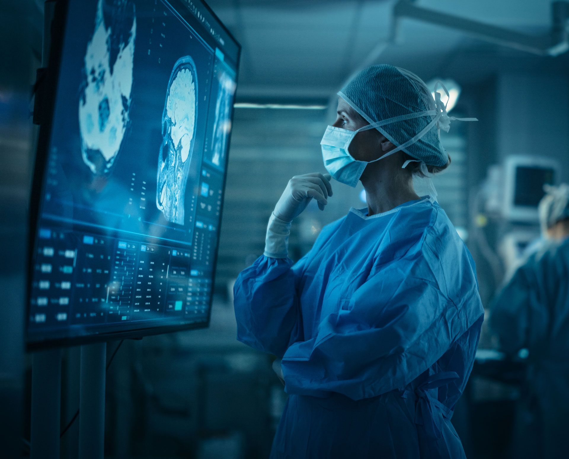 Surgeon in blue scrubs, looking at a screen displaying a brain scan in a surgical suite.