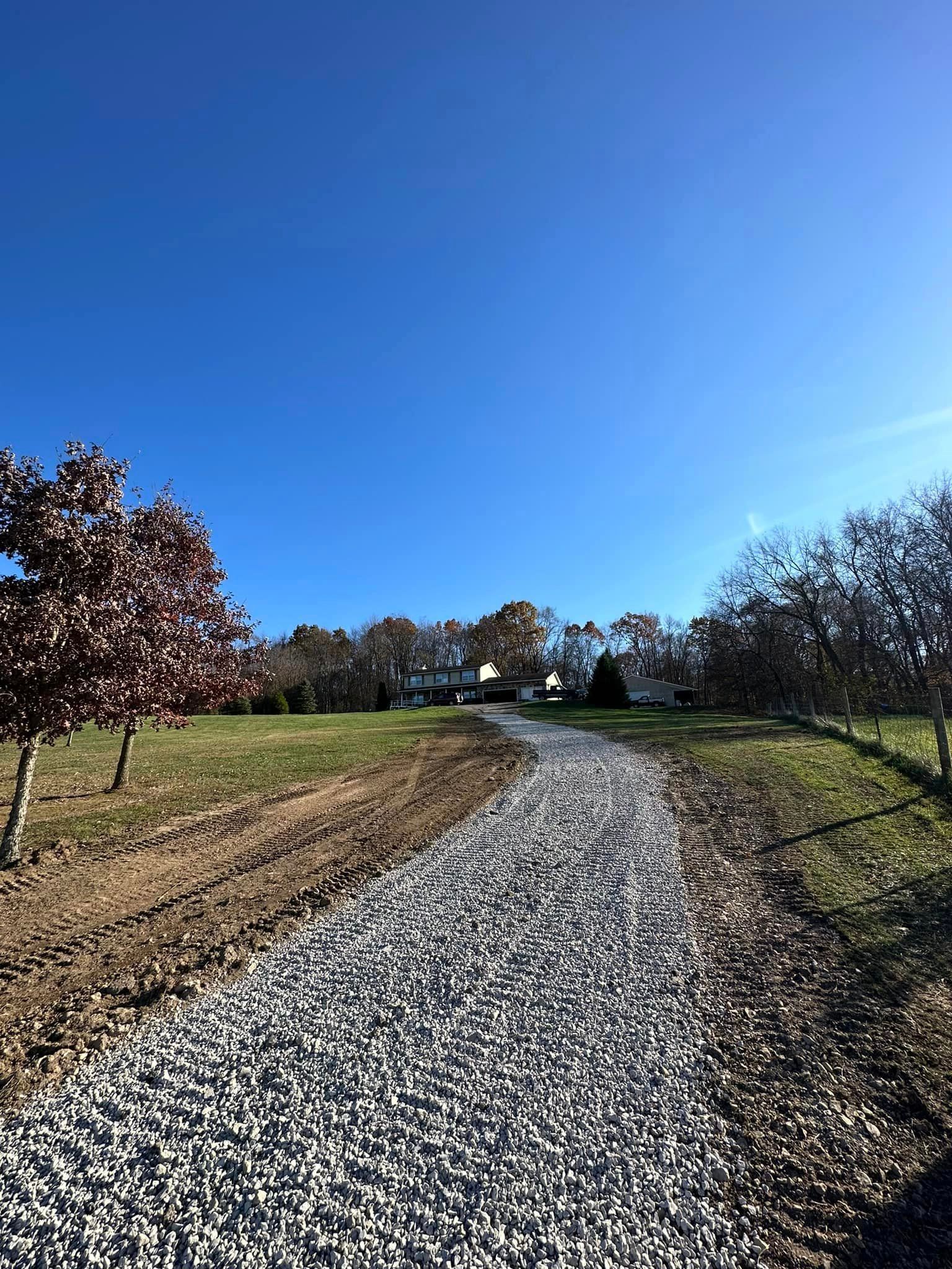 A gravel road going through a field with trees and a house in the background.