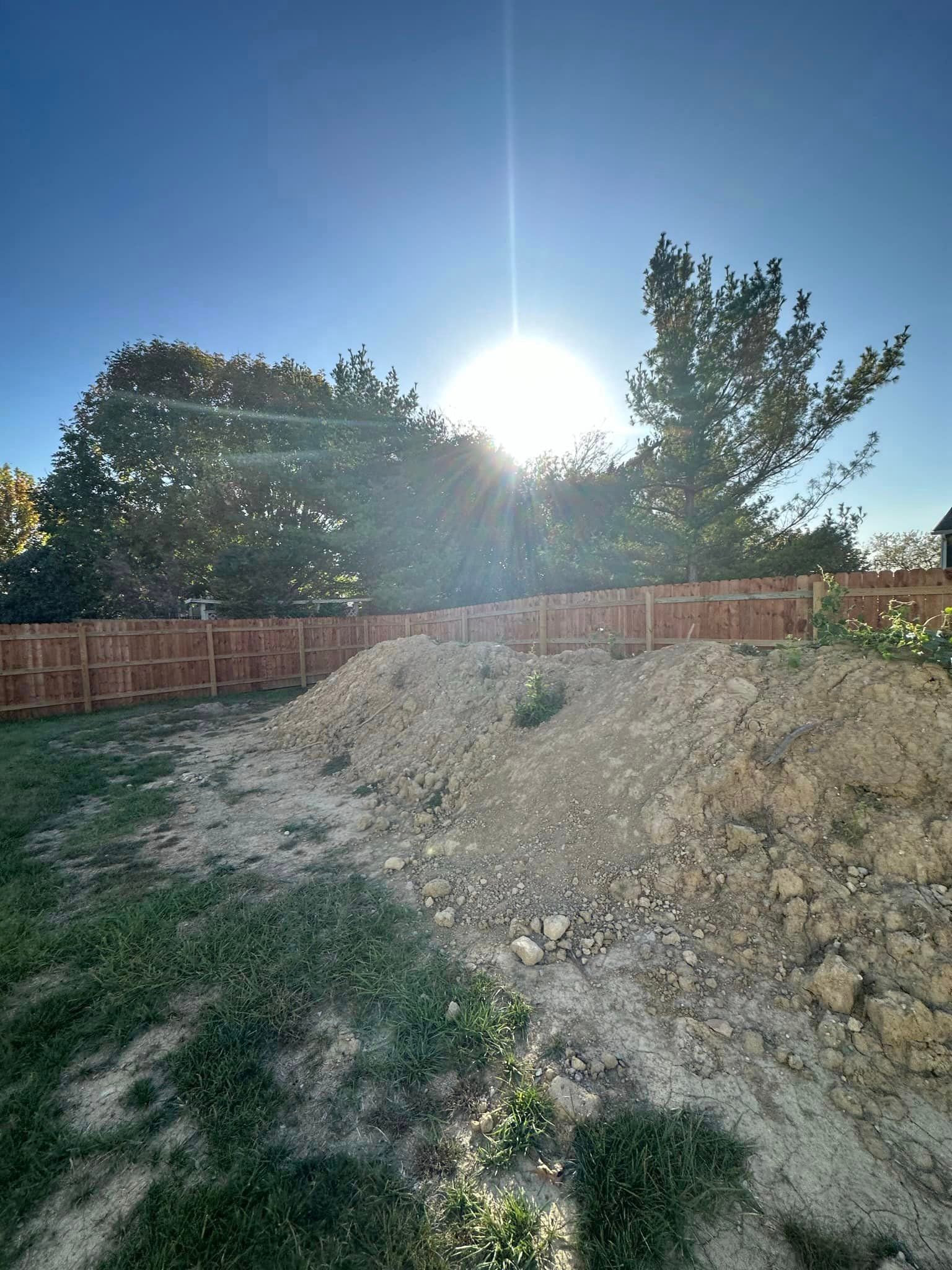 A pile of dirt in a backyard next to a wooden fence with the sun shining through the trees.