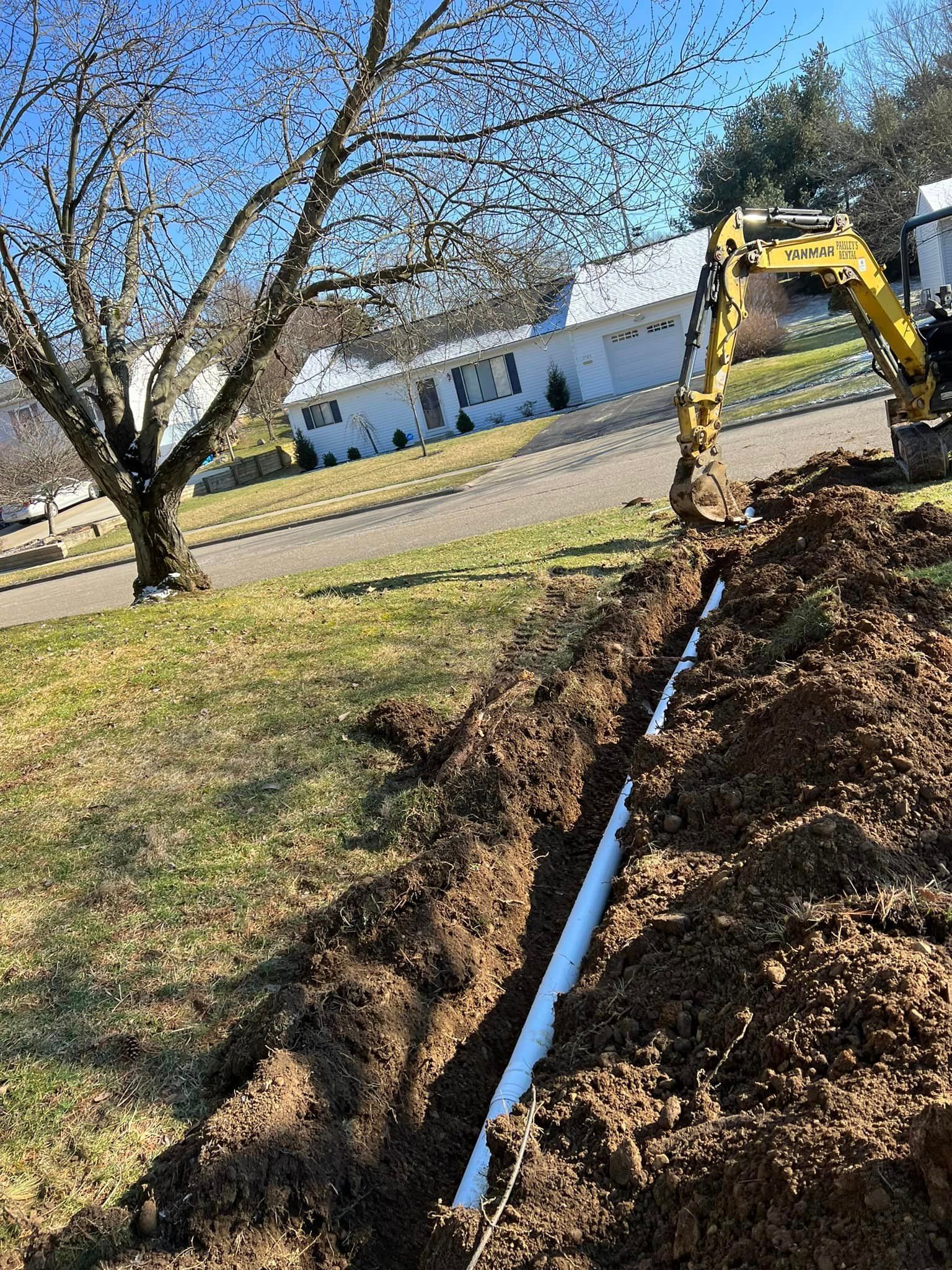 A yellow excavator is digging a trench in the dirt in front of a house.