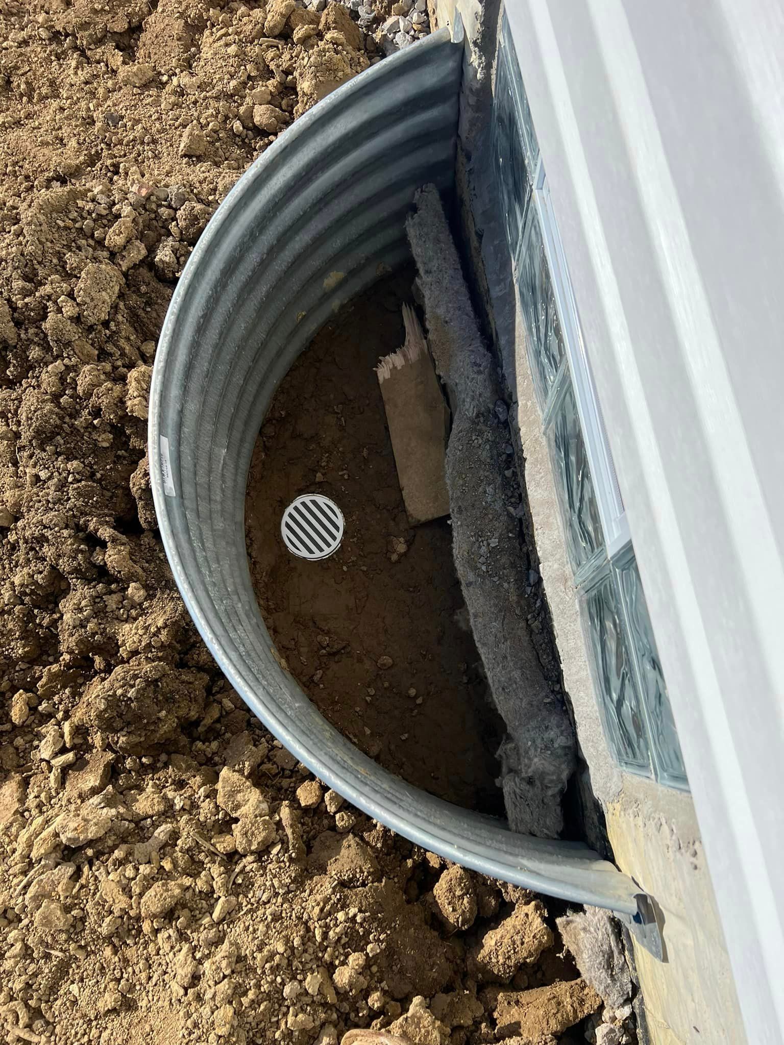 A metal window well is sitting in the dirt next to a house.