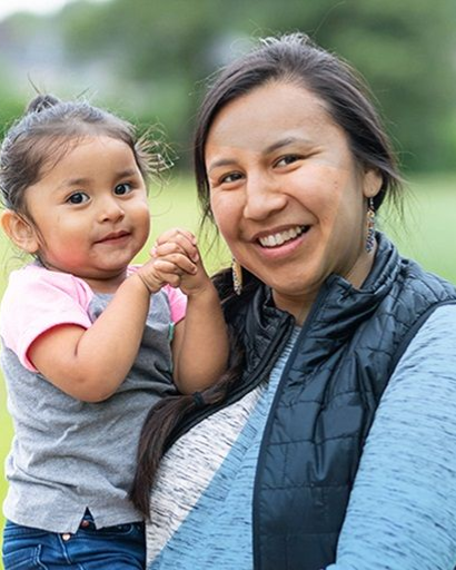 A smiling person holding a young child outdoors. Both are looking toward the camera in a grassy setting.