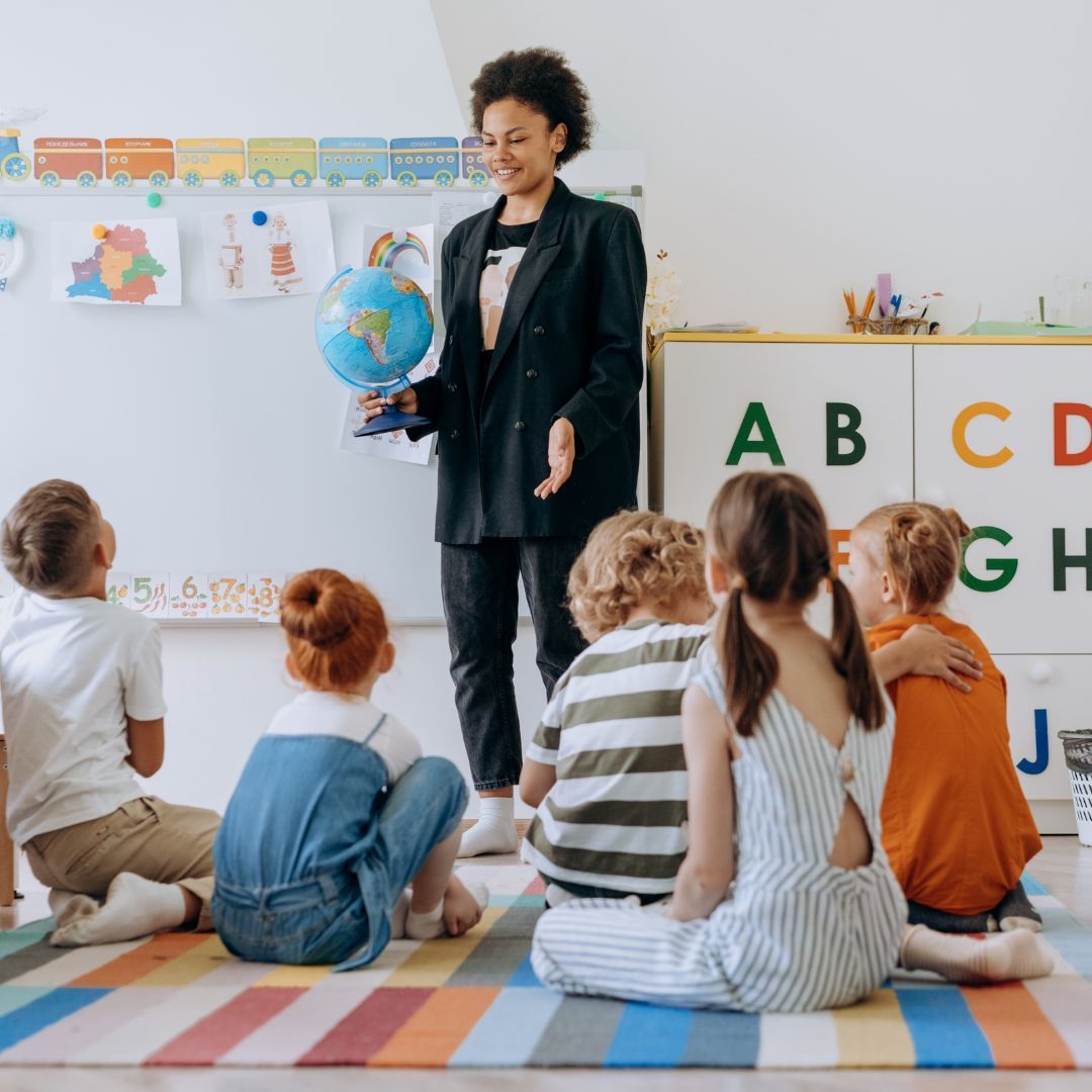 A teacher conducting a lesson while the students sit on a rug in front of her