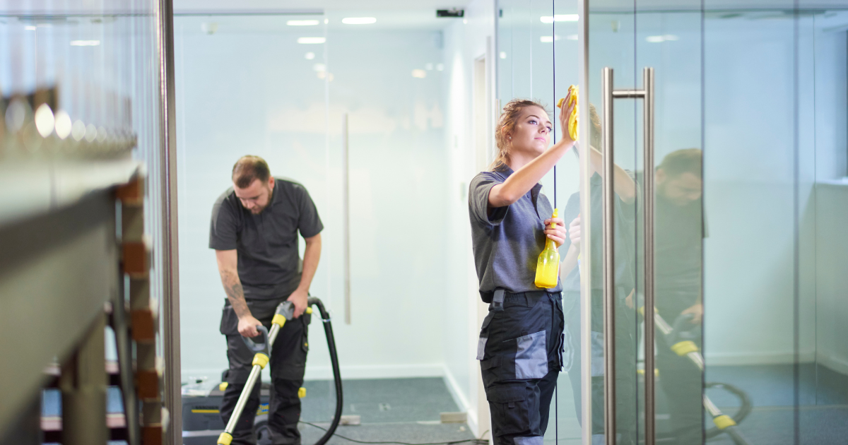 Two office cleaners cleaning a glass wall and vacuuming in an office hallway