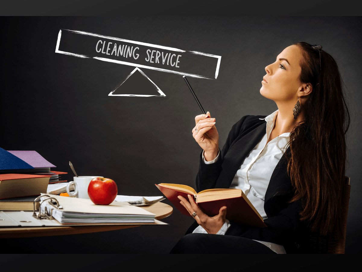 A school teacher sitting at a table crowded with books while looking up at a chalkboard drawing of a scale with the words [cleaning service] written along the side.