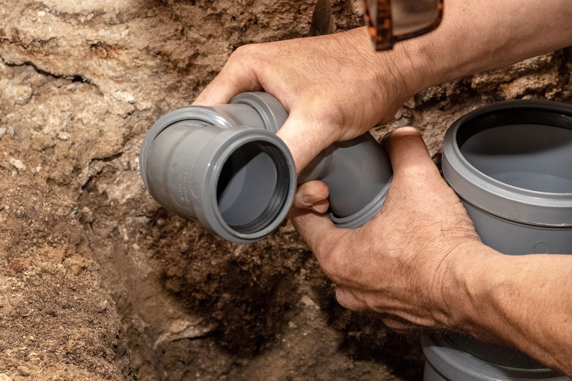 Hands connect gray PVC pipes in a dirt trench during plumbing installation