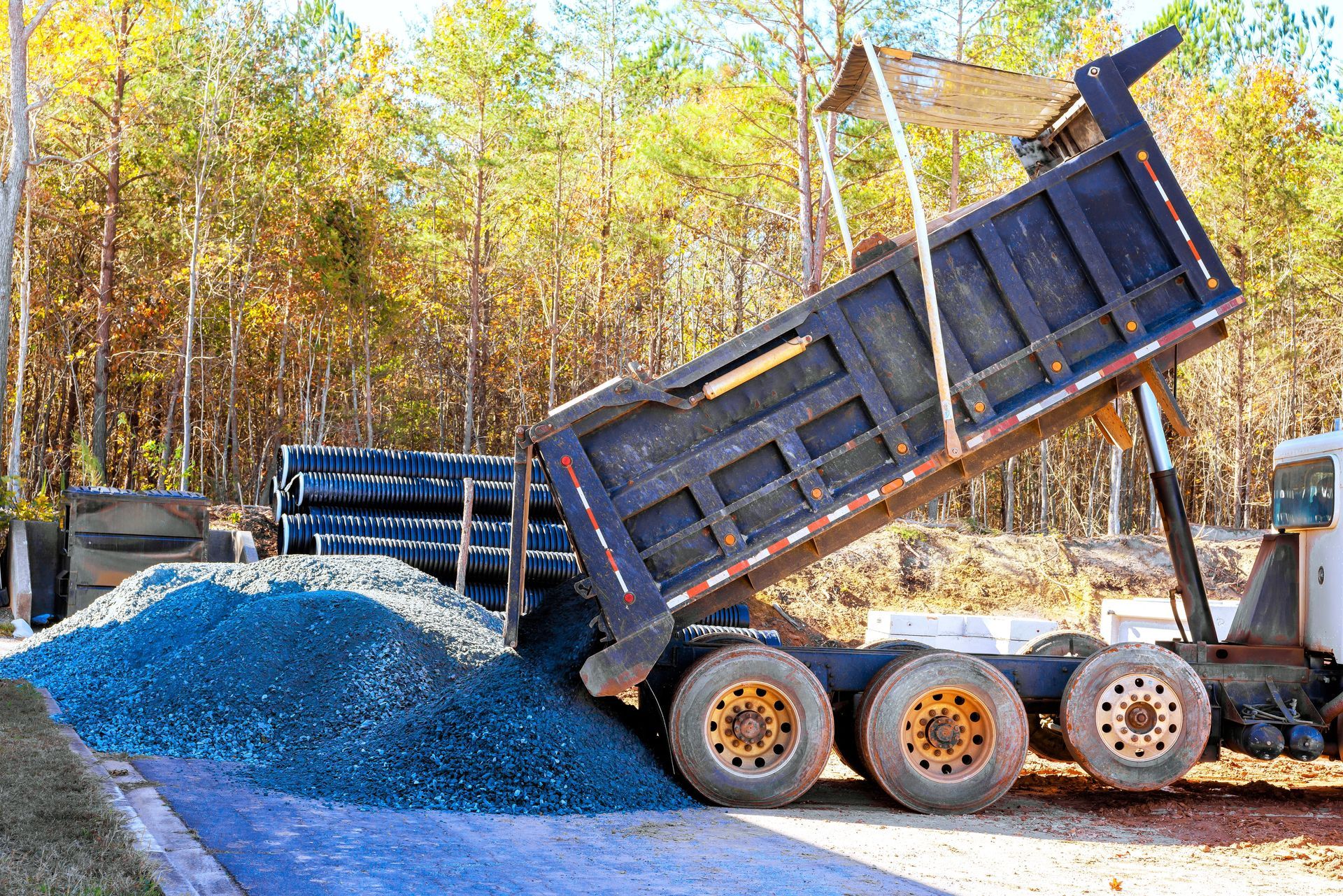 Dump truck unloading blue gravel at a construction site with autumn trees in the background