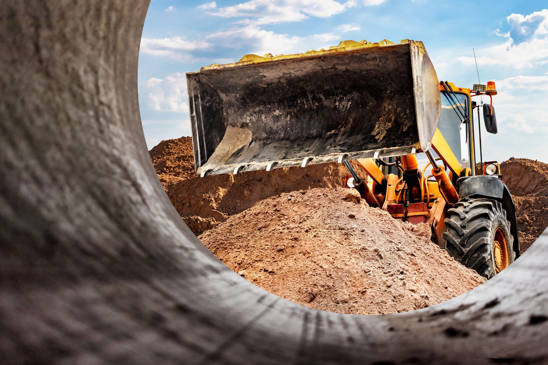 Yellow excavator dumping dirt at a construction site, viewed through a concrete pipe