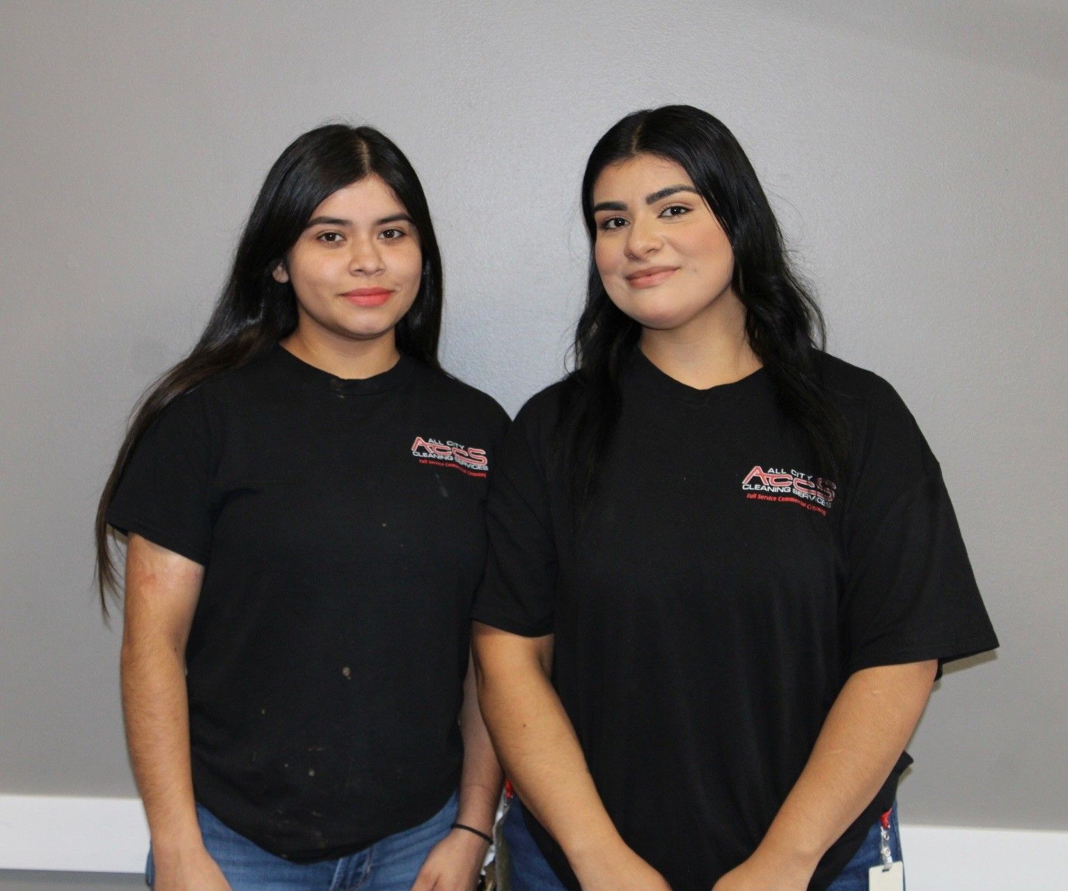 Two women in black shirts stand side-by-side, smiling against a gray wall.