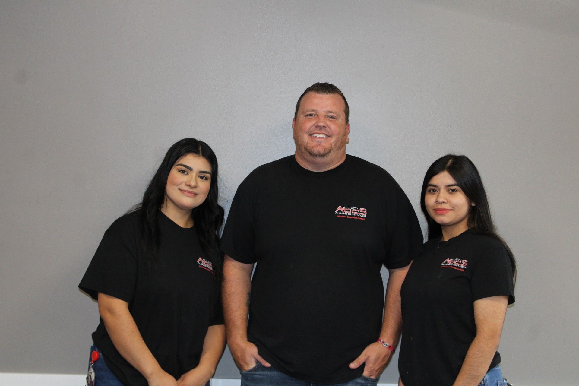 Three people in black t-shirts stand against a grey wall. A man in the center smiles, flanked by two women.
