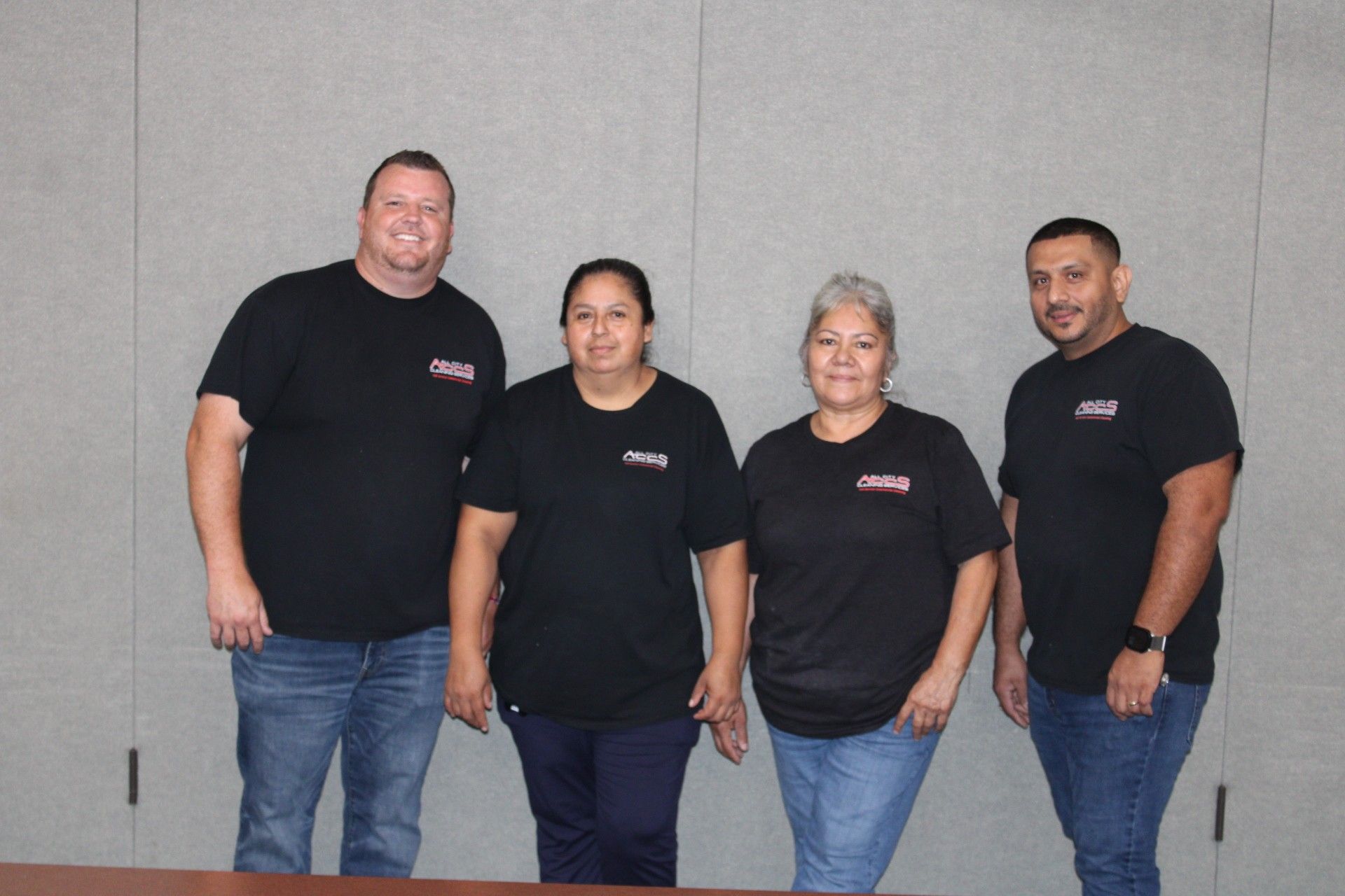 Four people in black shirts and jeans standing in front of a gray wall.