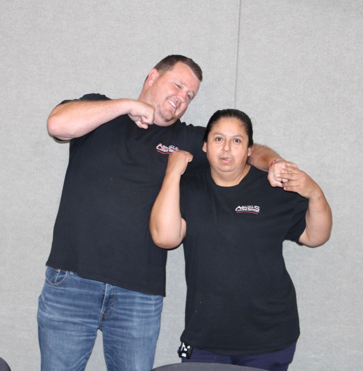 Man and woman in black shirts, making flexing poses. Light background.