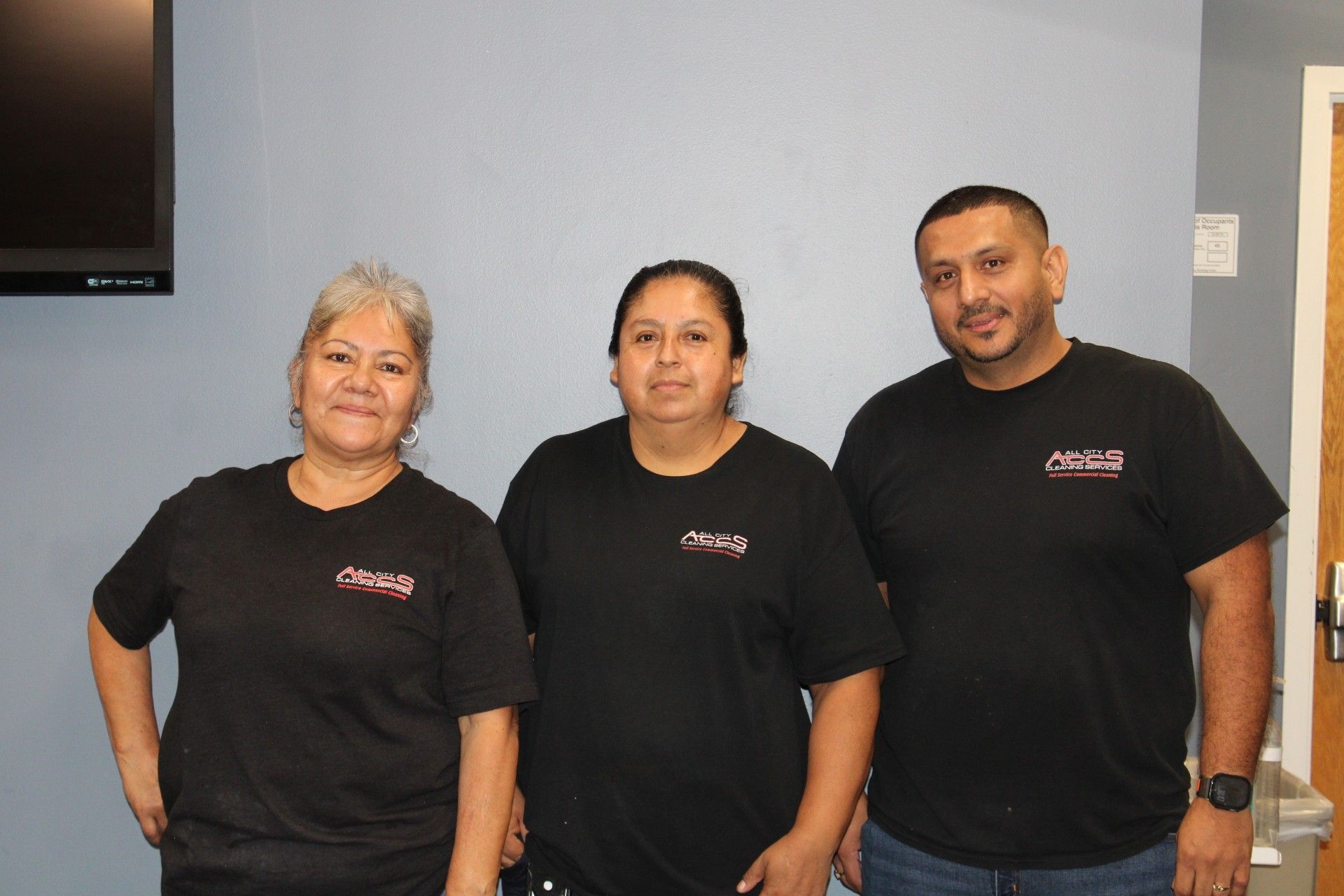 Three people in black shirts stand indoors, smiling.