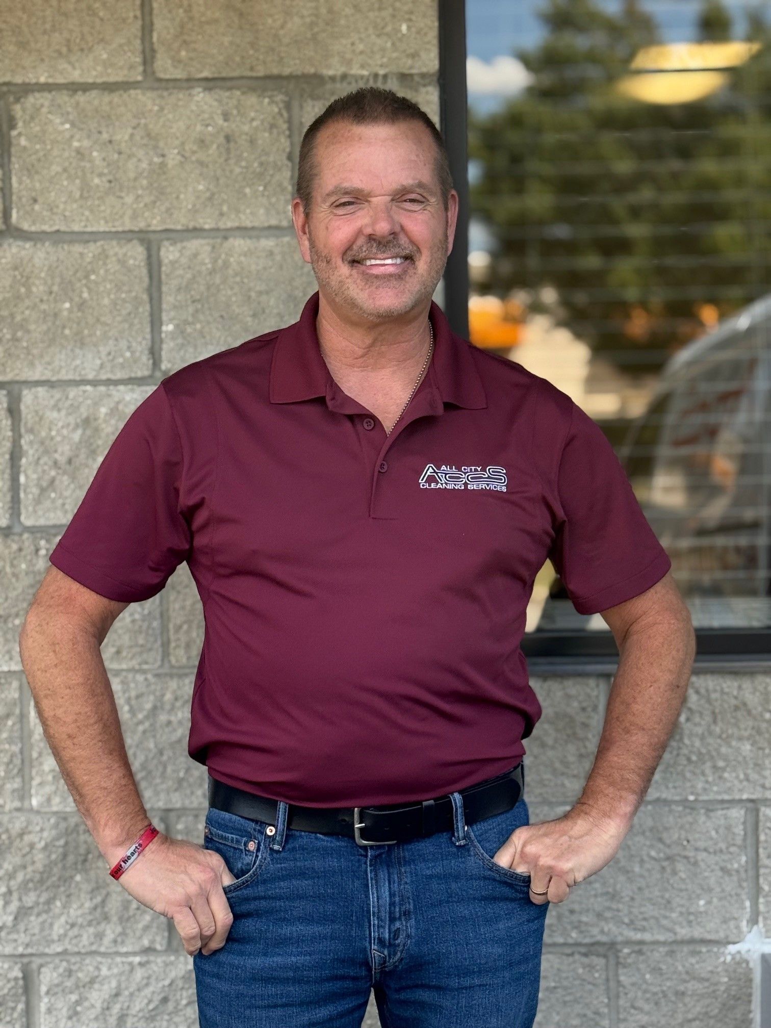 Man in maroon polo shirt and jeans smiles, hands in pockets, in front of a brick wall.