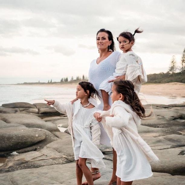 A woman is carrying two little girls on her shoulders on a rocky beach.