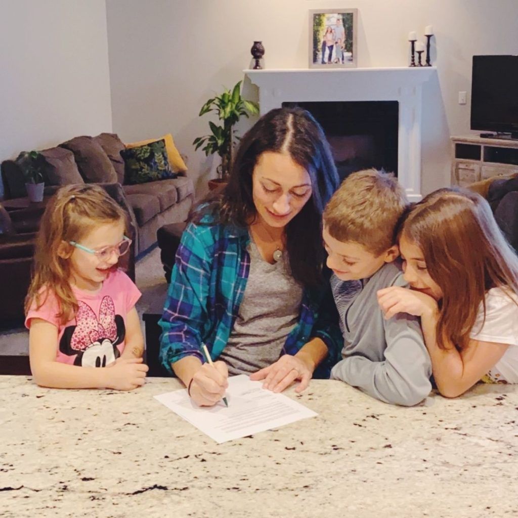 A woman is sitting at a table with three children and writing on a piece of paper