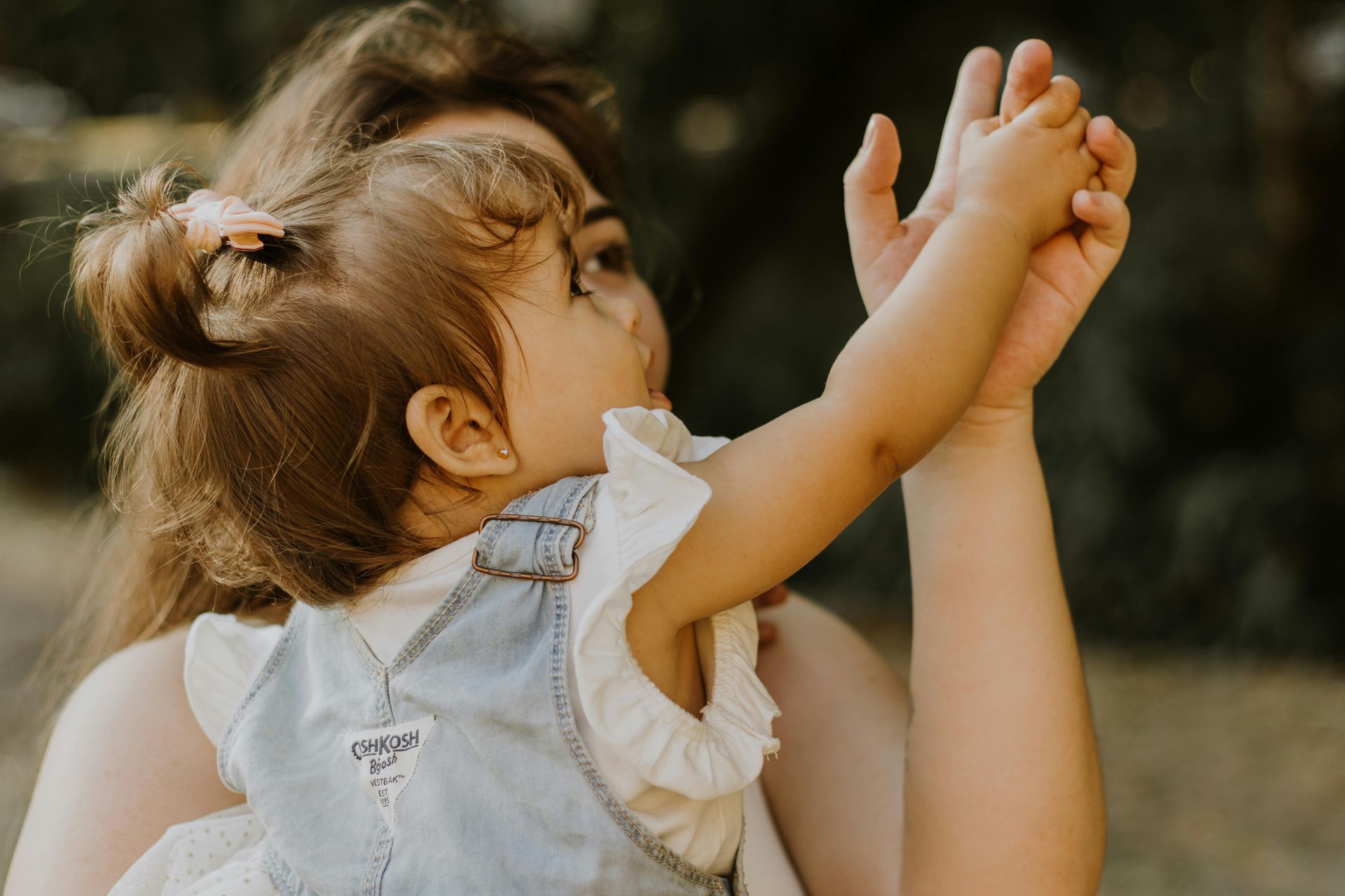 Mother holding a baby girl, arms raised. Baby wears denim overalls, looking up.