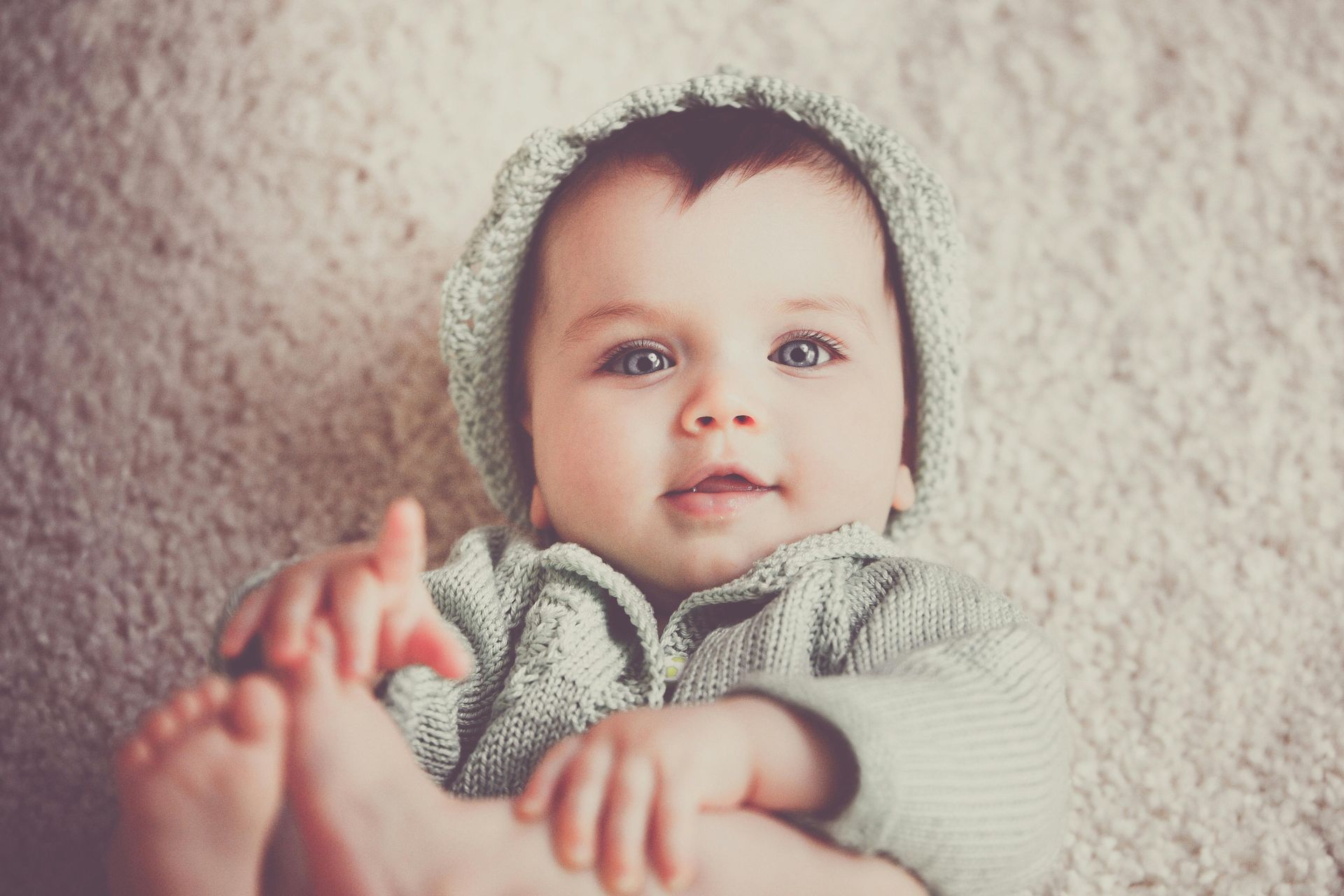 Baby with light eyes wearing a green knit hat and sweater, lying on a fluffy white rug.