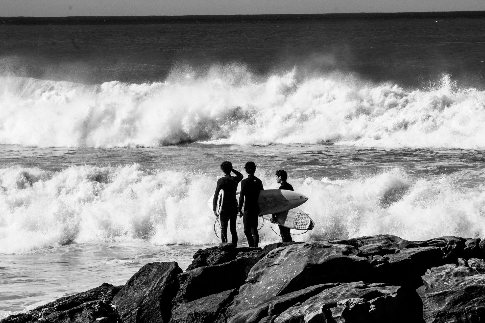Three surfers are standing on rocks near the ocean looking at the waves.