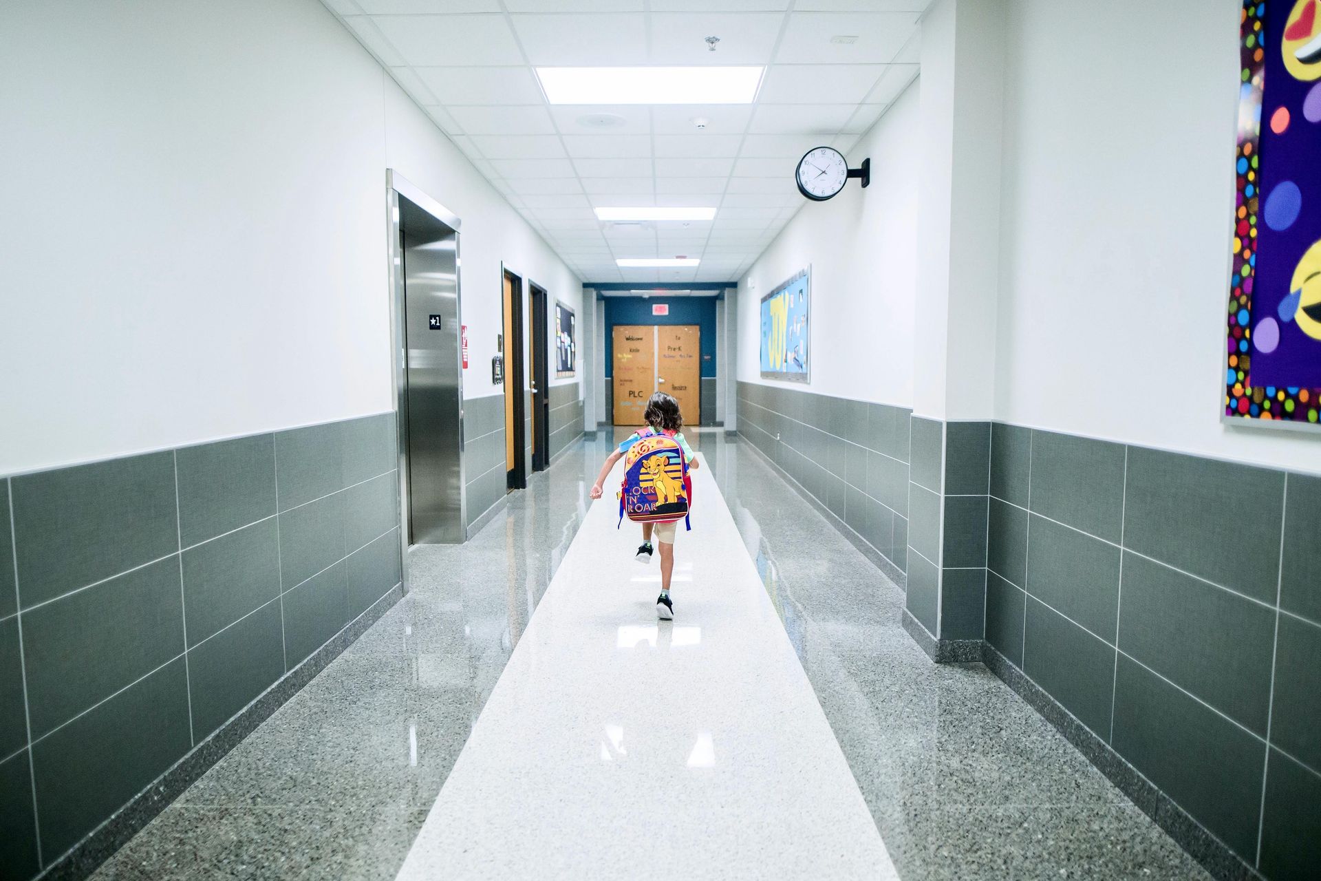 Child running down a school hallway, wearing a backpack, with gray and white walls and flooring.