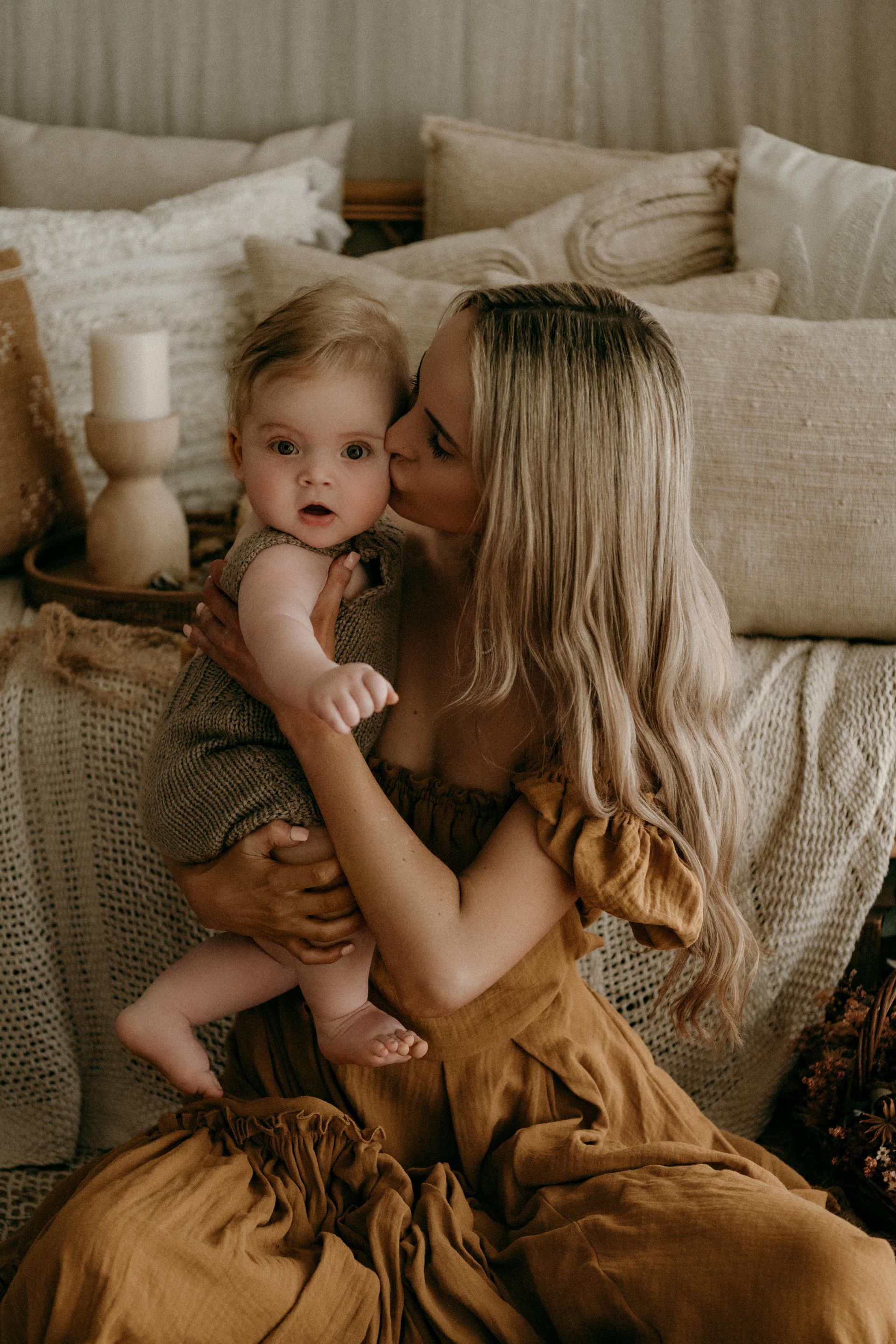Woman in a mustard dress kisses a baby's cheek. They sit surrounded by beige pillows on a bed.