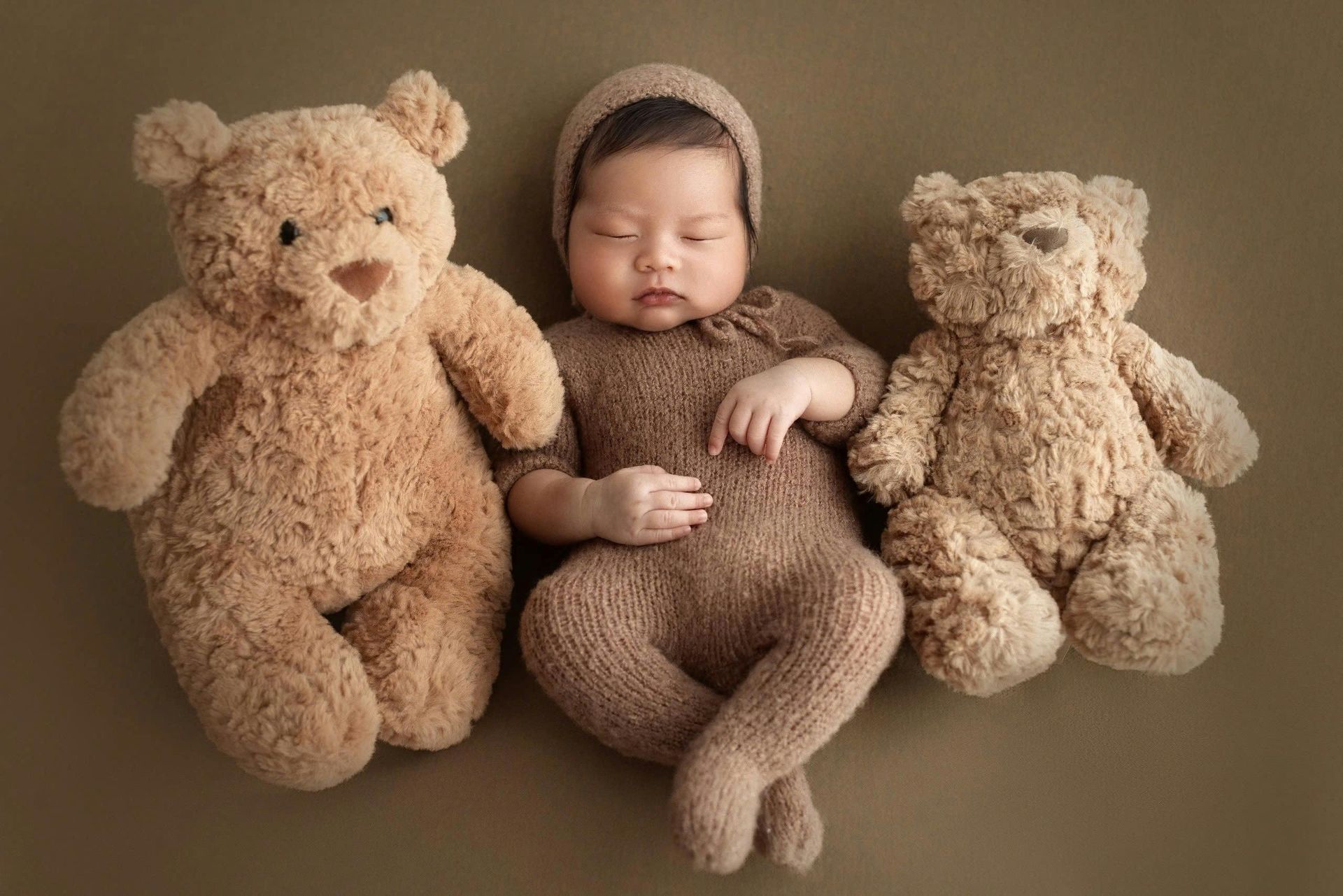 Newborn baby wearing brown outfit and hat, sleeping between two teddy bears on a brown surface.
