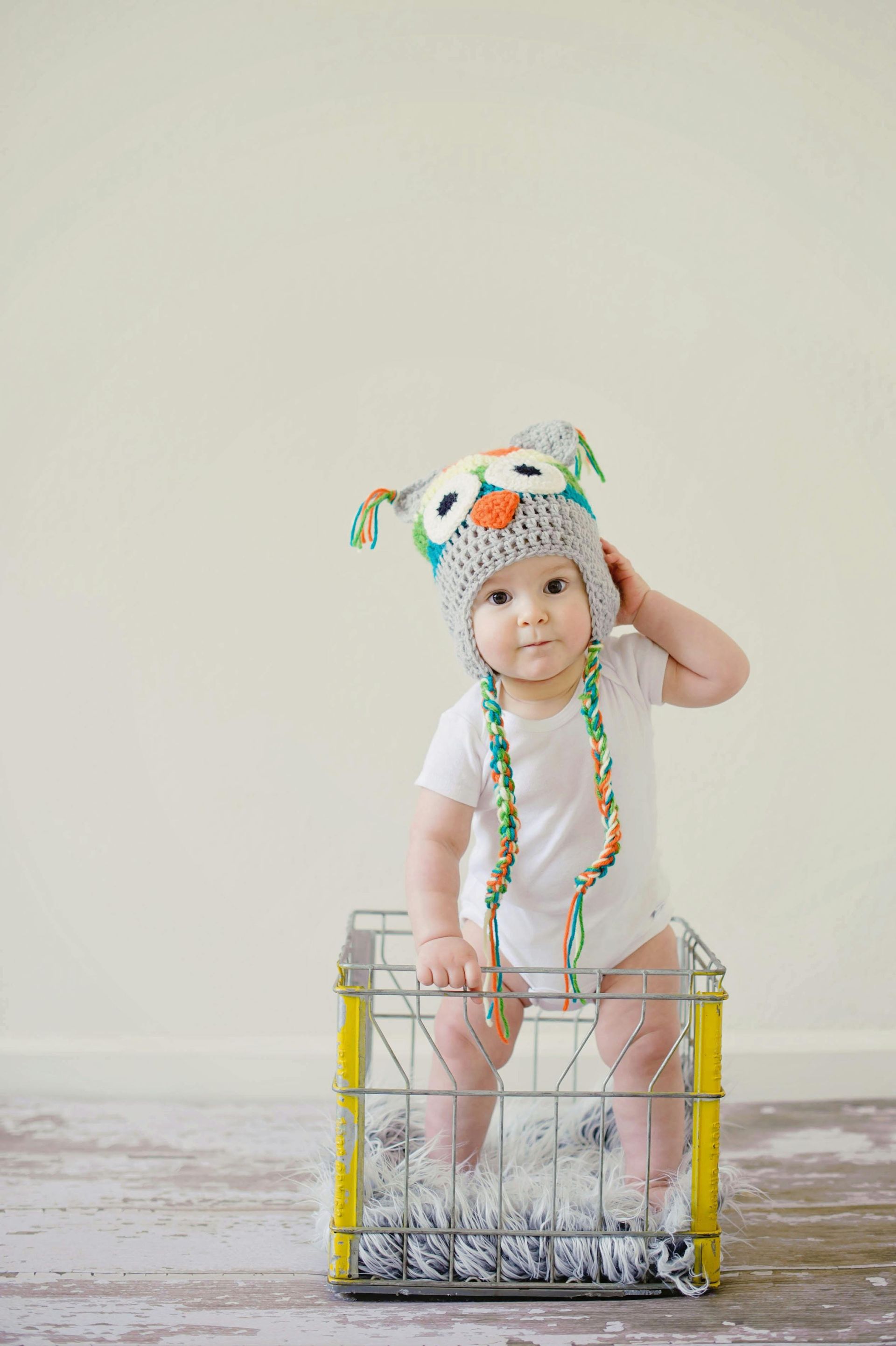 Baby in owl hat stands in yellow crate, hand on head, looking at camera. White onesie, colorful beads.