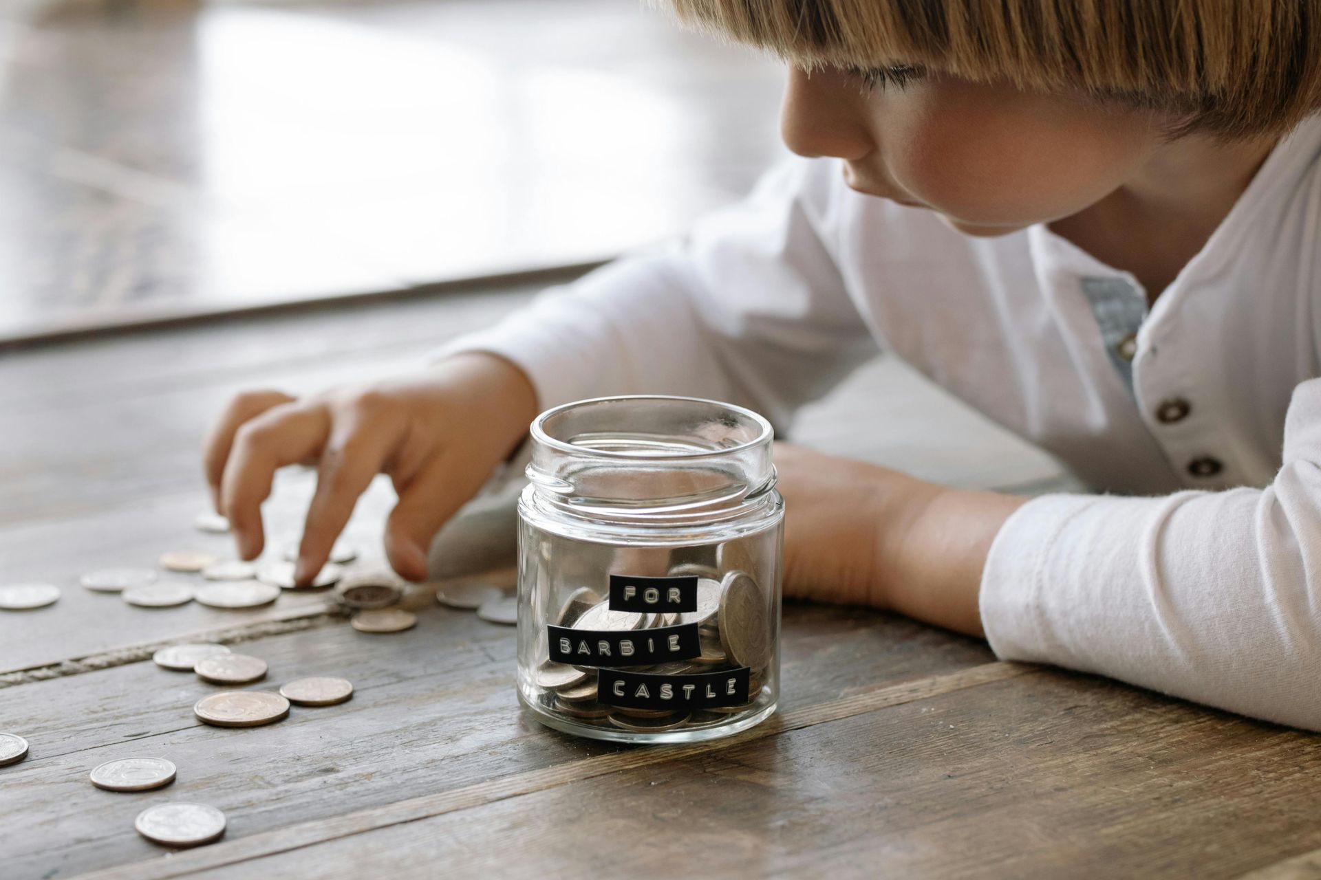 A young boy is putting coins into a jar on a wooden table.