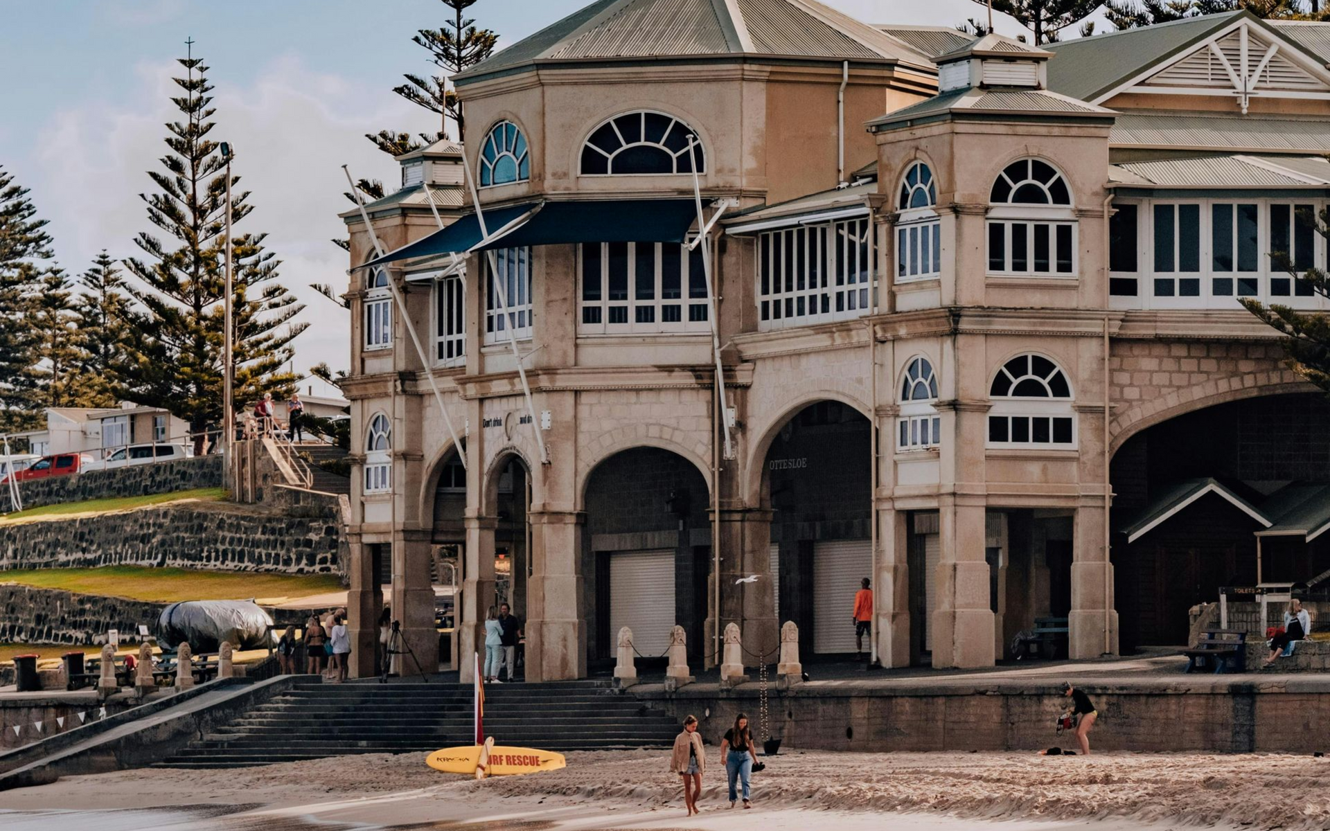 Beachside building with arched entrance; two people walk along the shore, waves crash.