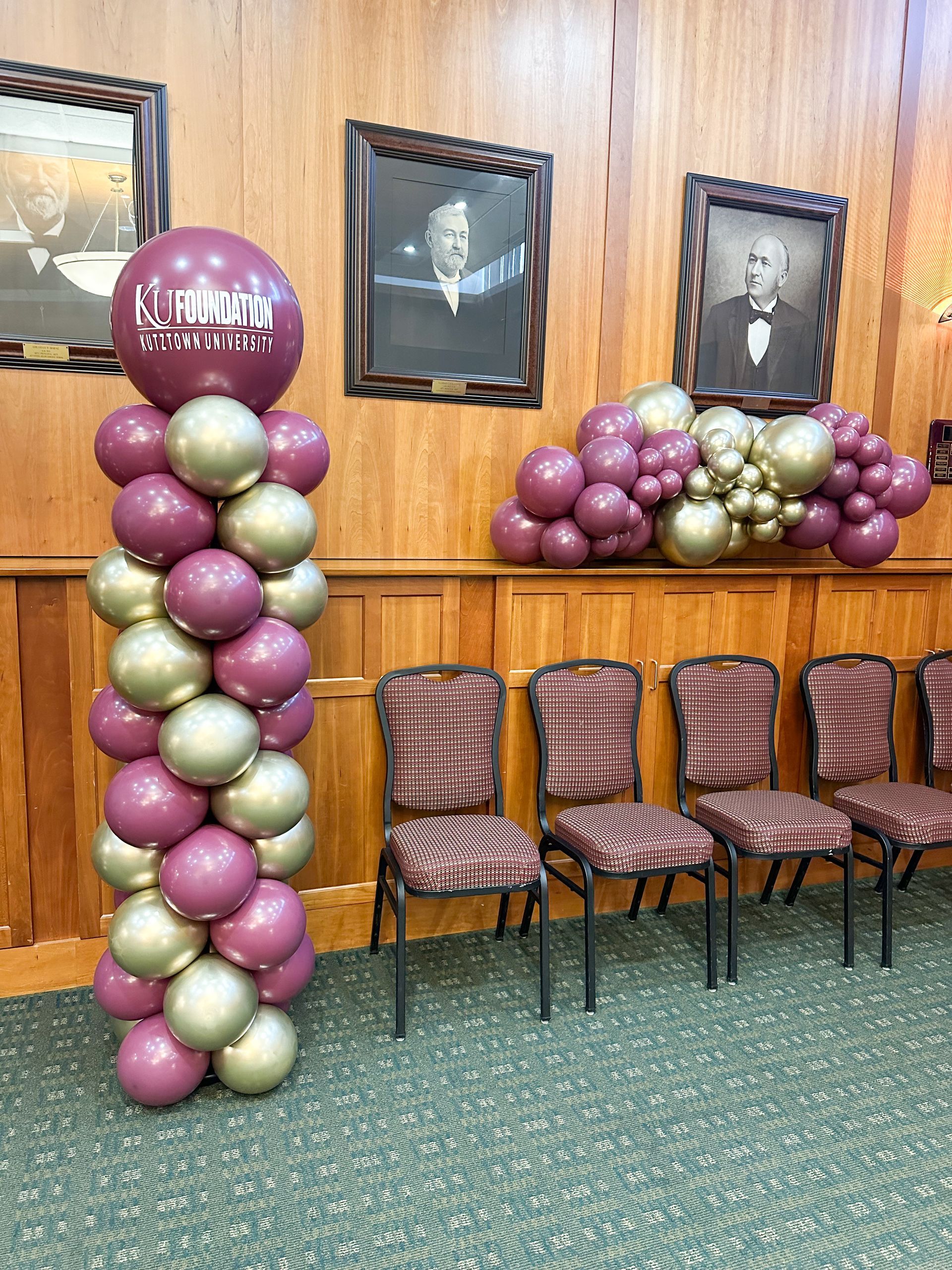 Balloon decorations and chairs in front of wood-paneled wall with framed portraits.