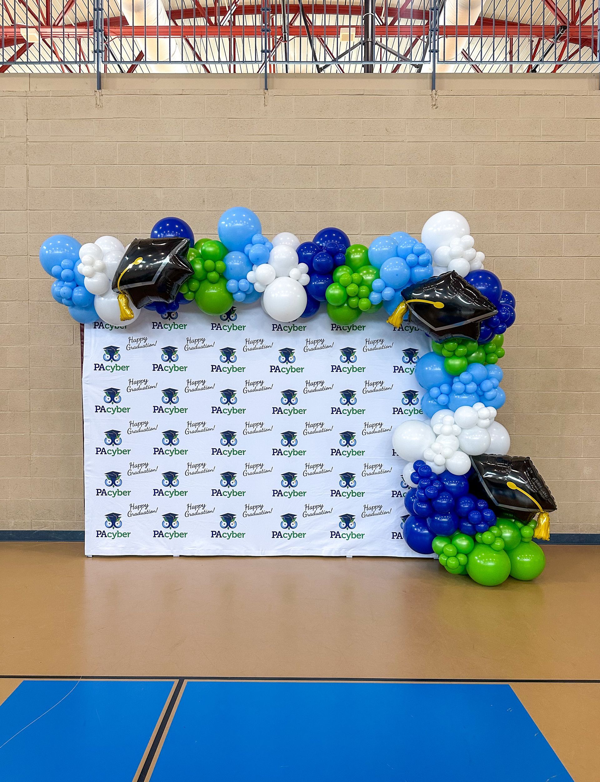 Graduation backdrop with blue, white, green, and black balloons, mortarboards, and a branded banner, on a gymnasium floor.