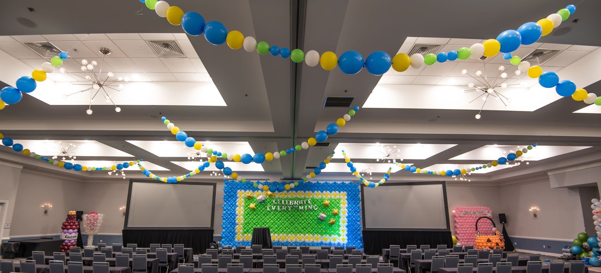 Ballroom decorated with blue, yellow, and green balloons for a celebration. Stage setup with screens and seating.