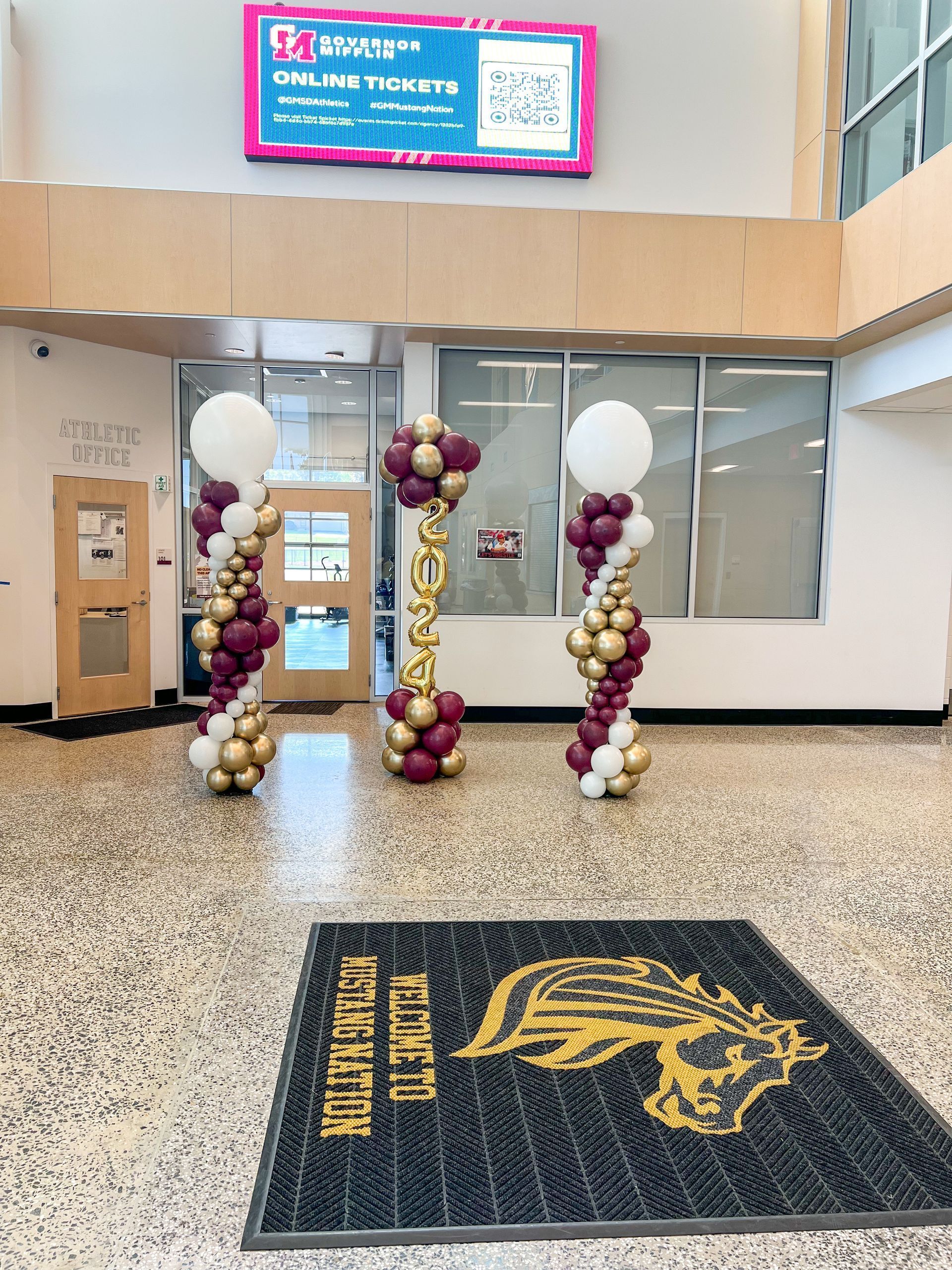 Entrance to a building with balloon decorations in burgundy, gold, and white. A welcome mat is in front.