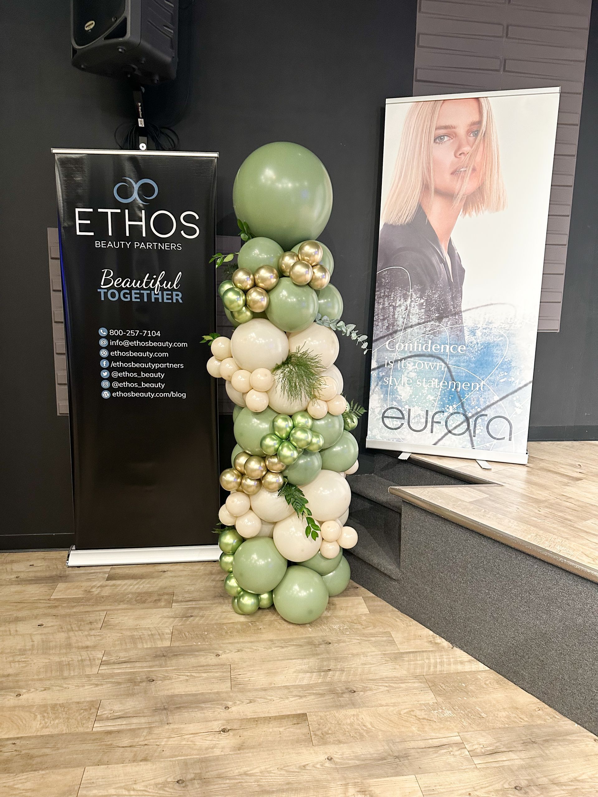 Event setup: balloon column, banners. Earth tone balloons: green, gold, white. ETHOS and Eufara banners on stage.