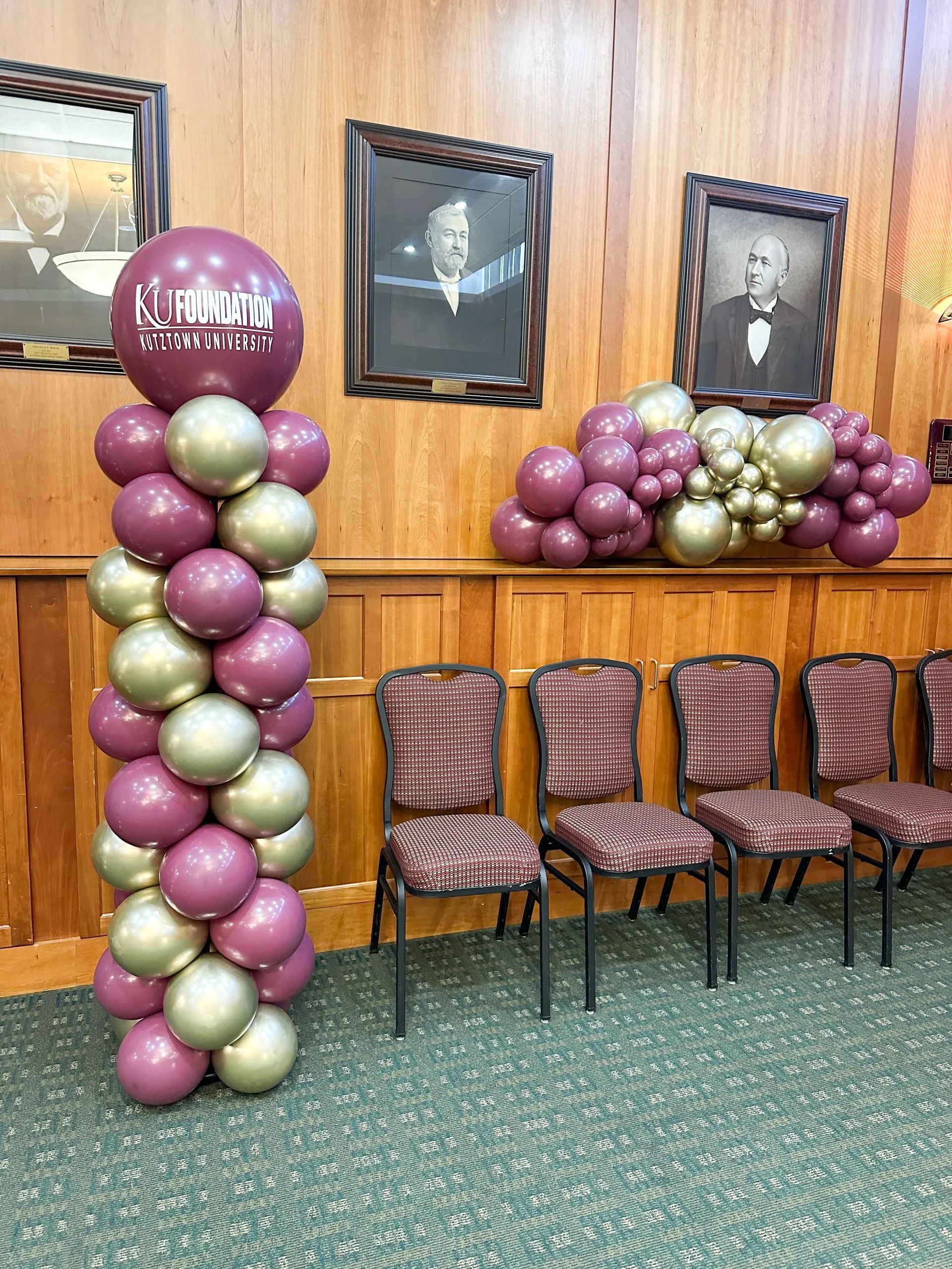 Balloon decorations with chairs in front of wood-paneled wall, featuring portraits. Purple and gold balloons.