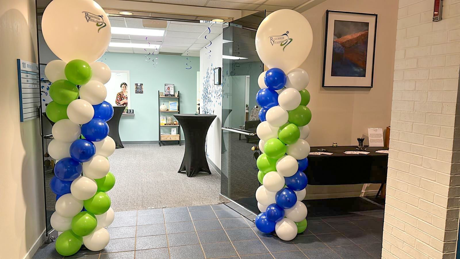 Two balloon columns, white, blue, and green, flank a doorway. A white balloon tops each column.