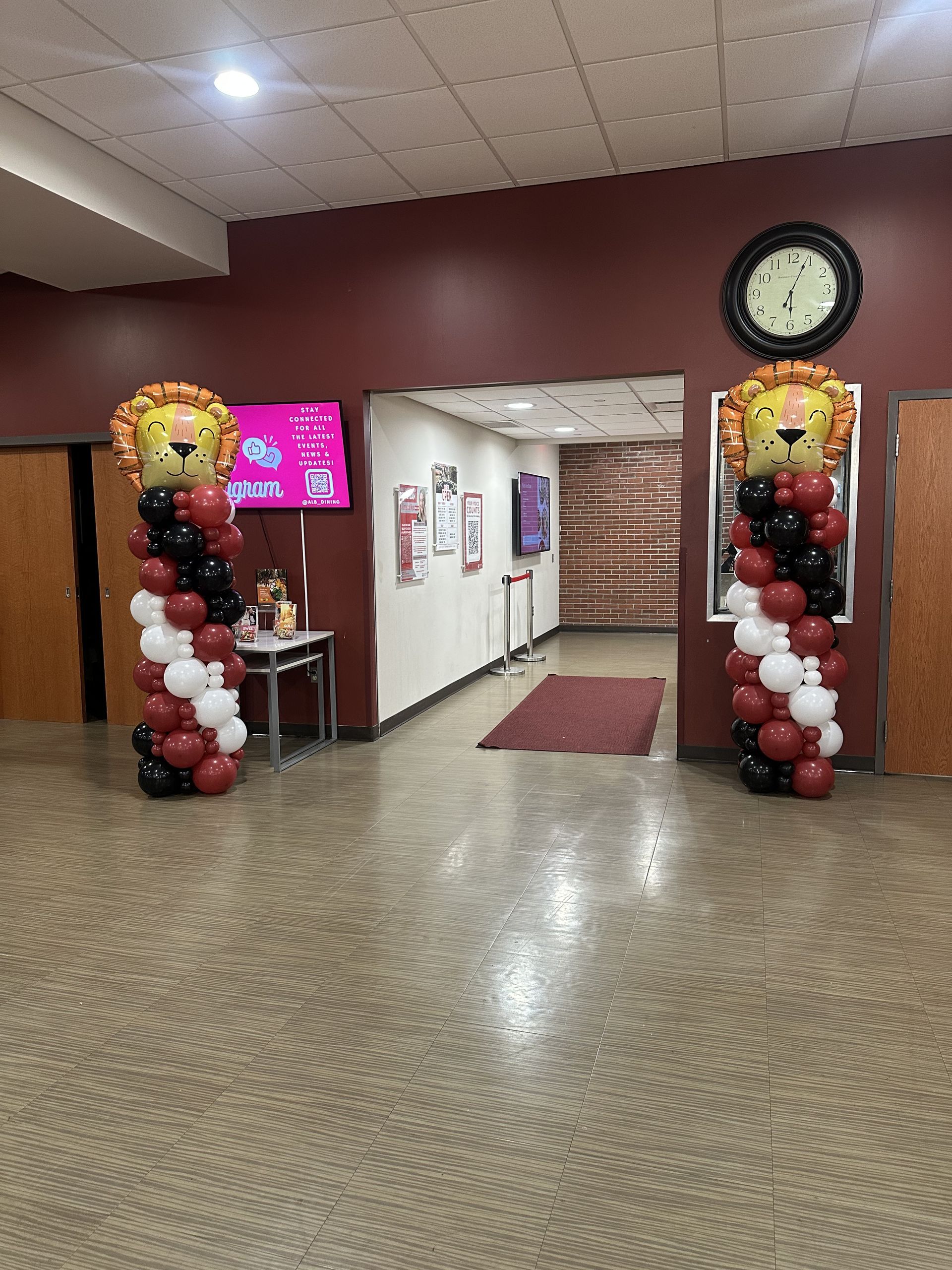 Entrance with balloon columns topped with lion faces, red/black/white balloons. Hallway with clock, door, and screens.
