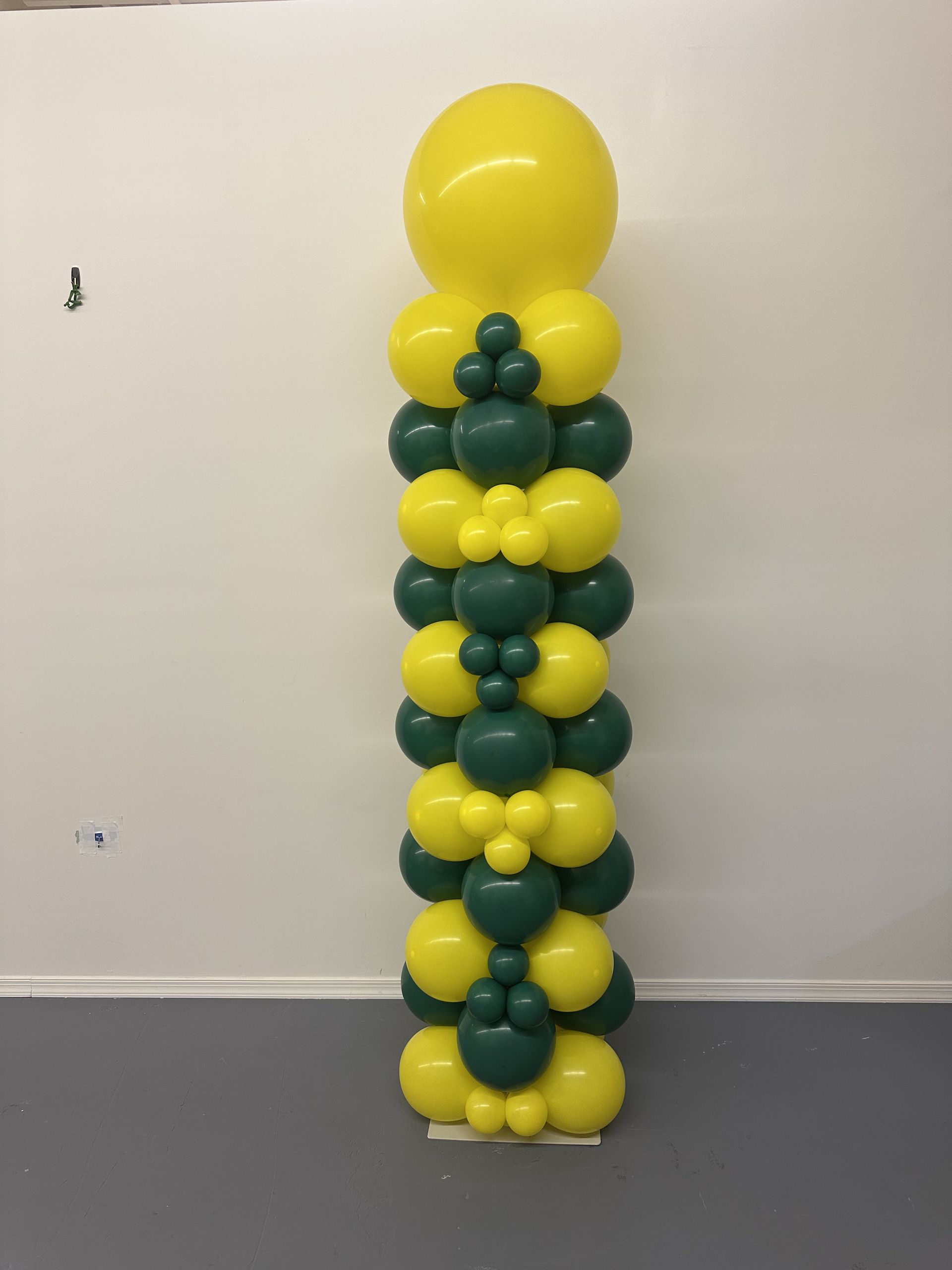 Yellow and green balloon column with a large yellow balloon on top, against a white wall and gray floor.