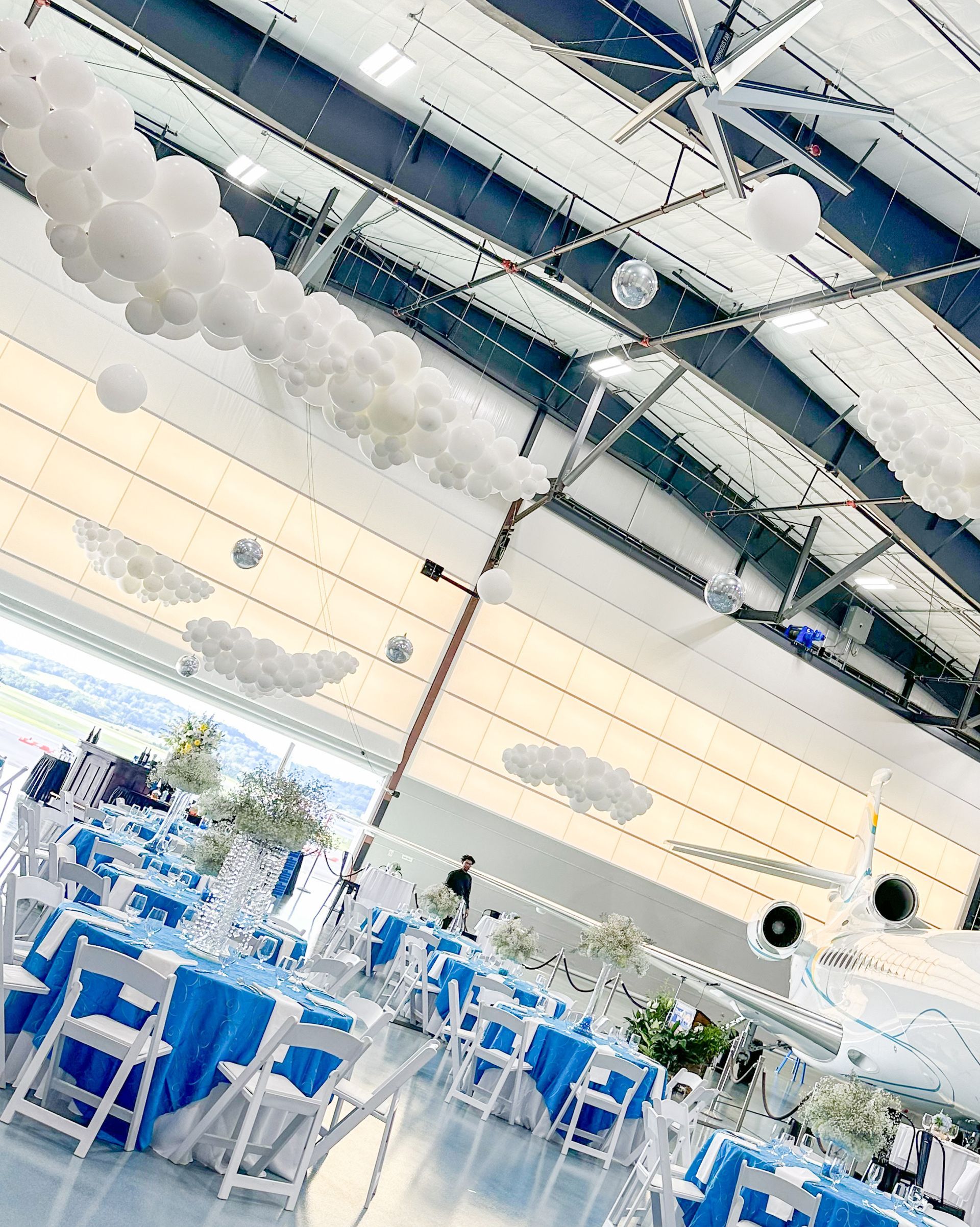 Blue-tabled event decor in large, high-ceilinged space with cloud decorations and plane, white chairs, and glass.