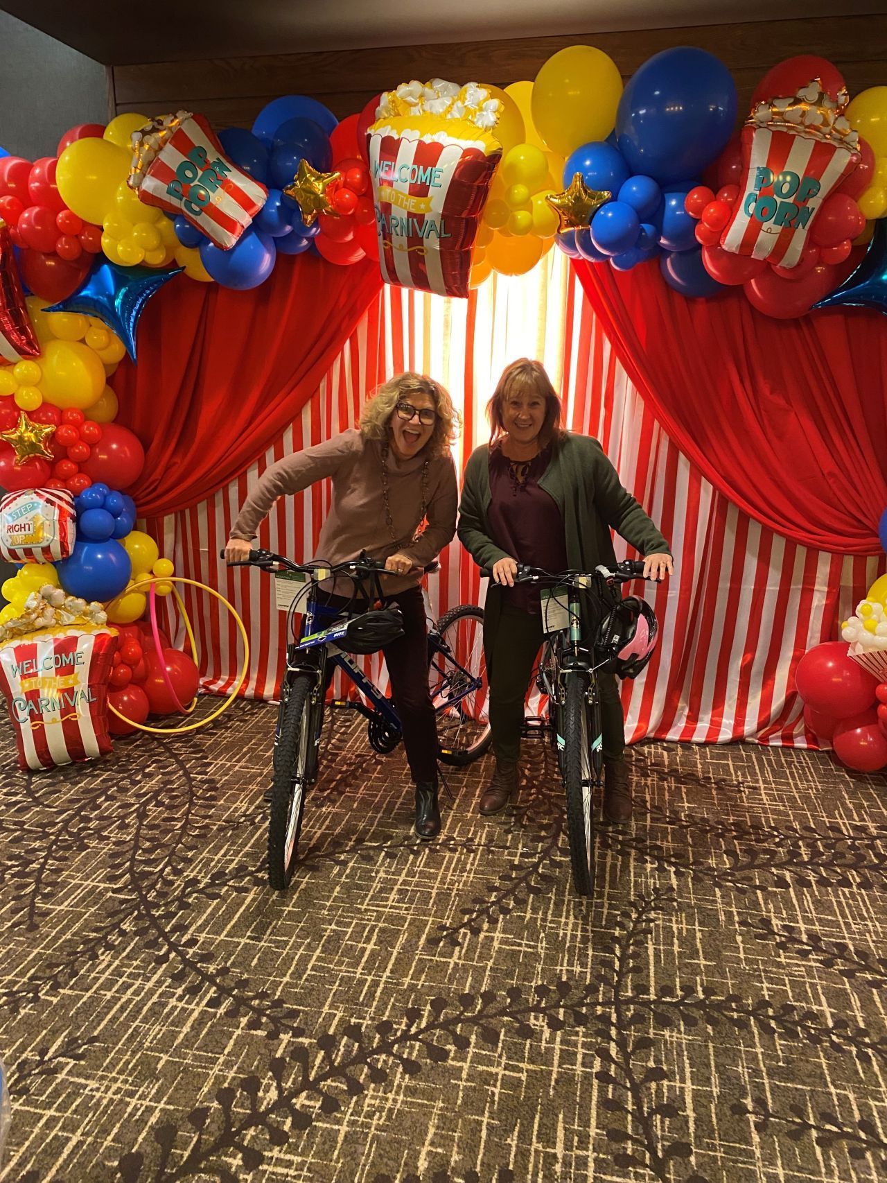 Two women on bikes, smiling in front of a circus-themed backdrop with balloons, popcorn, and red curtains.